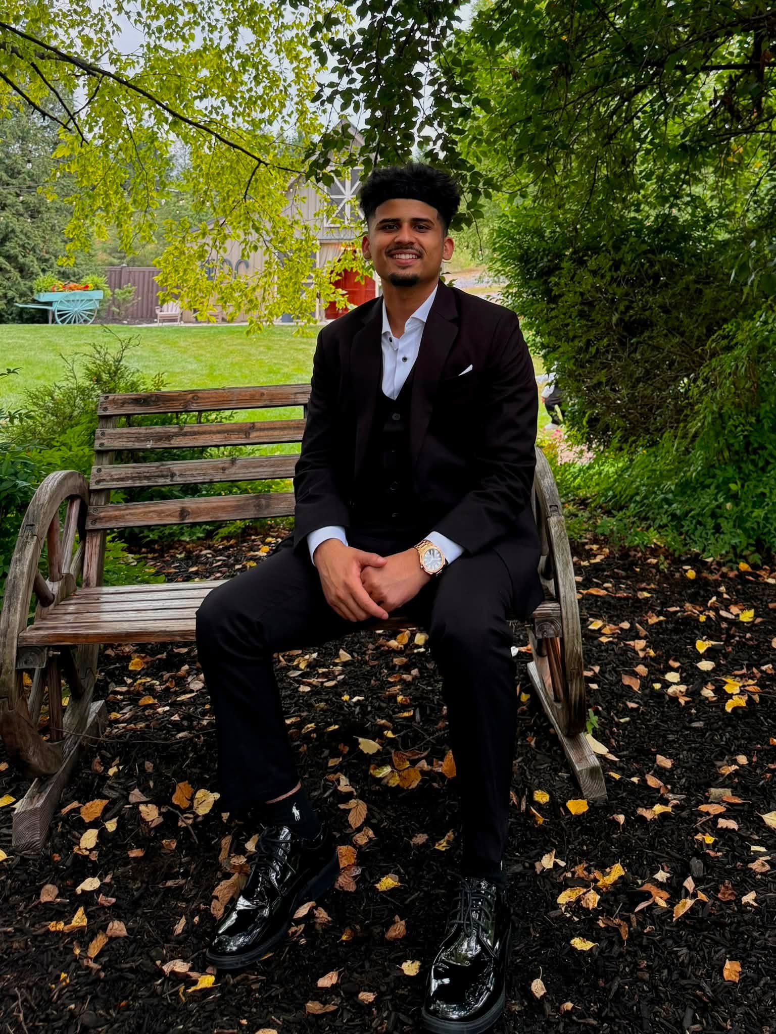 Young man in formal suit sitting on a park bench surrounded by green trees and fallen leaves.