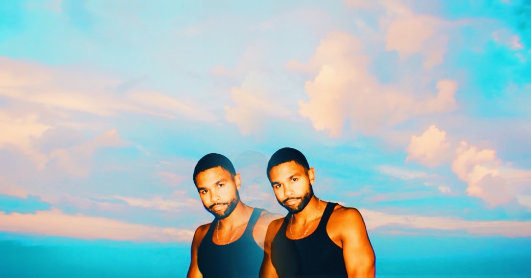 Two men with short hair and beards wearing sleeveless shirts stand outdoors against a sky with clouds.