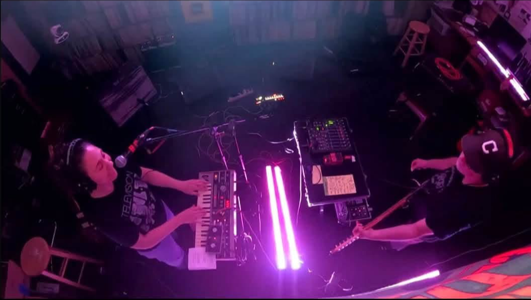 Two musicians perform on stage with electronic equipment, illuminated by pink and purple lights, viewed from above.