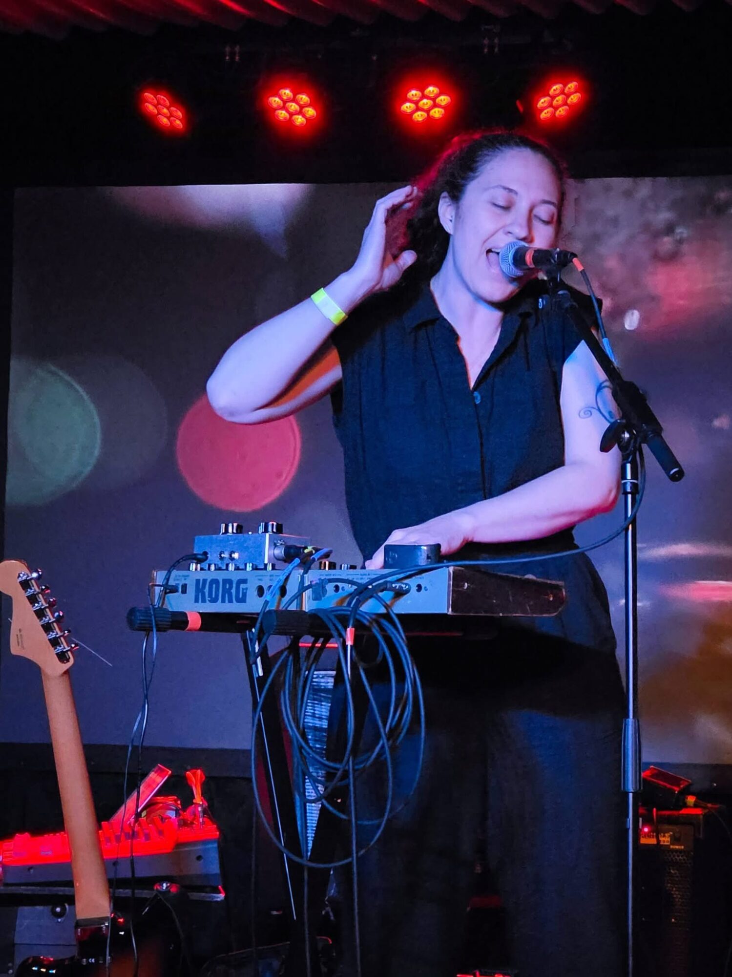 Female singer with long hair, wearing a dark shirt, performing with electronic music equipment on stage, microphone in hand.