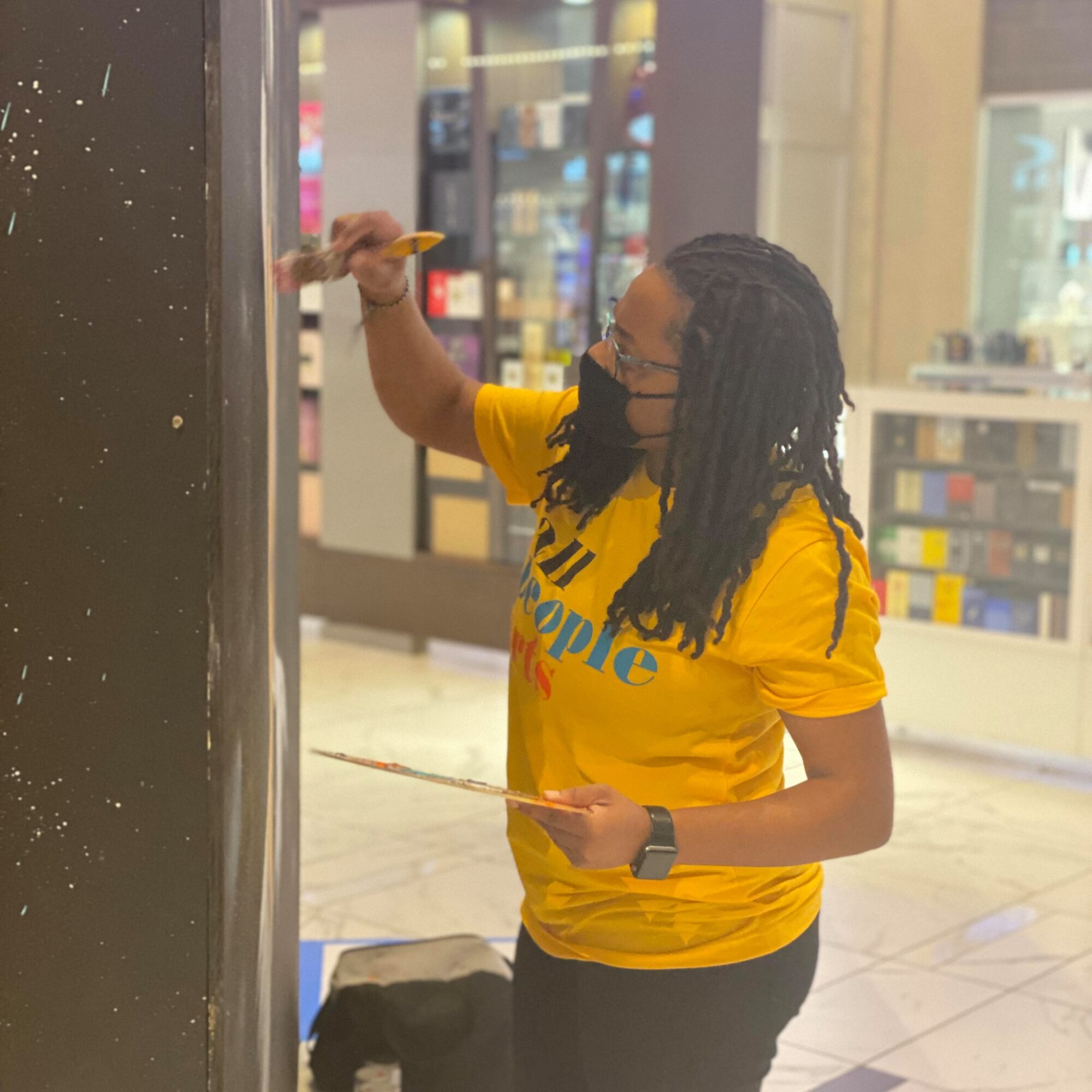 Person with long dreadlocks wearing a yellow shirt and black mask, writing on a black pillar inside a shopping mall.