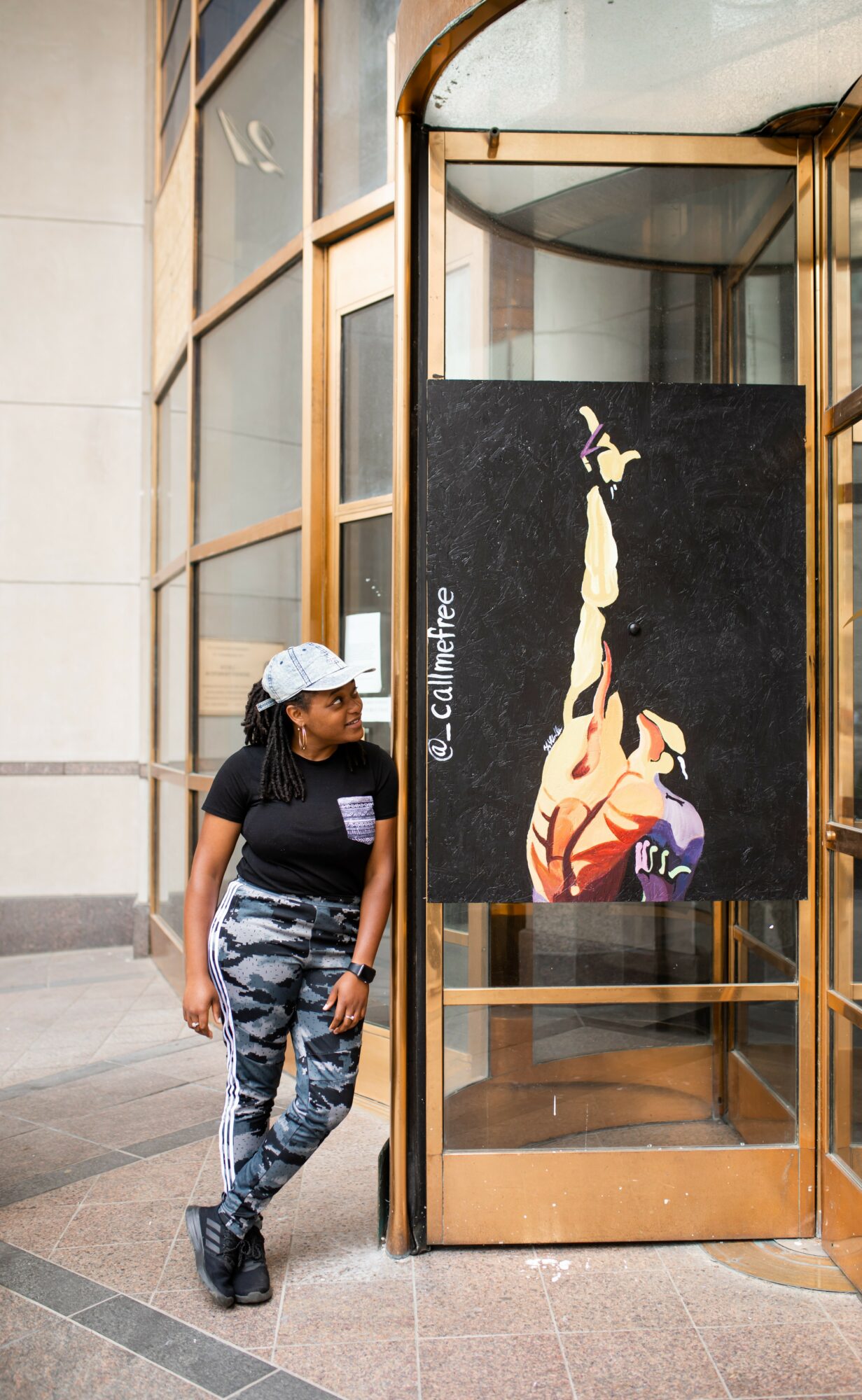 Young girl wearing a white cap, black shirt, and camouflage pants stands next to a glass display case with artwork inside.