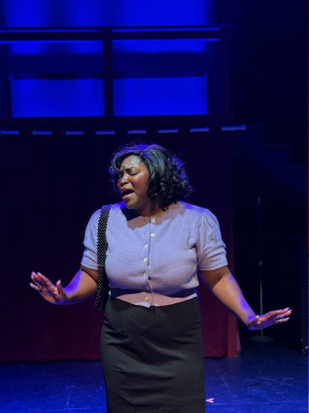 Woman singing on stage with dark background, wearing a light-colored top and dark skirt, expressive face, arms outstretched.