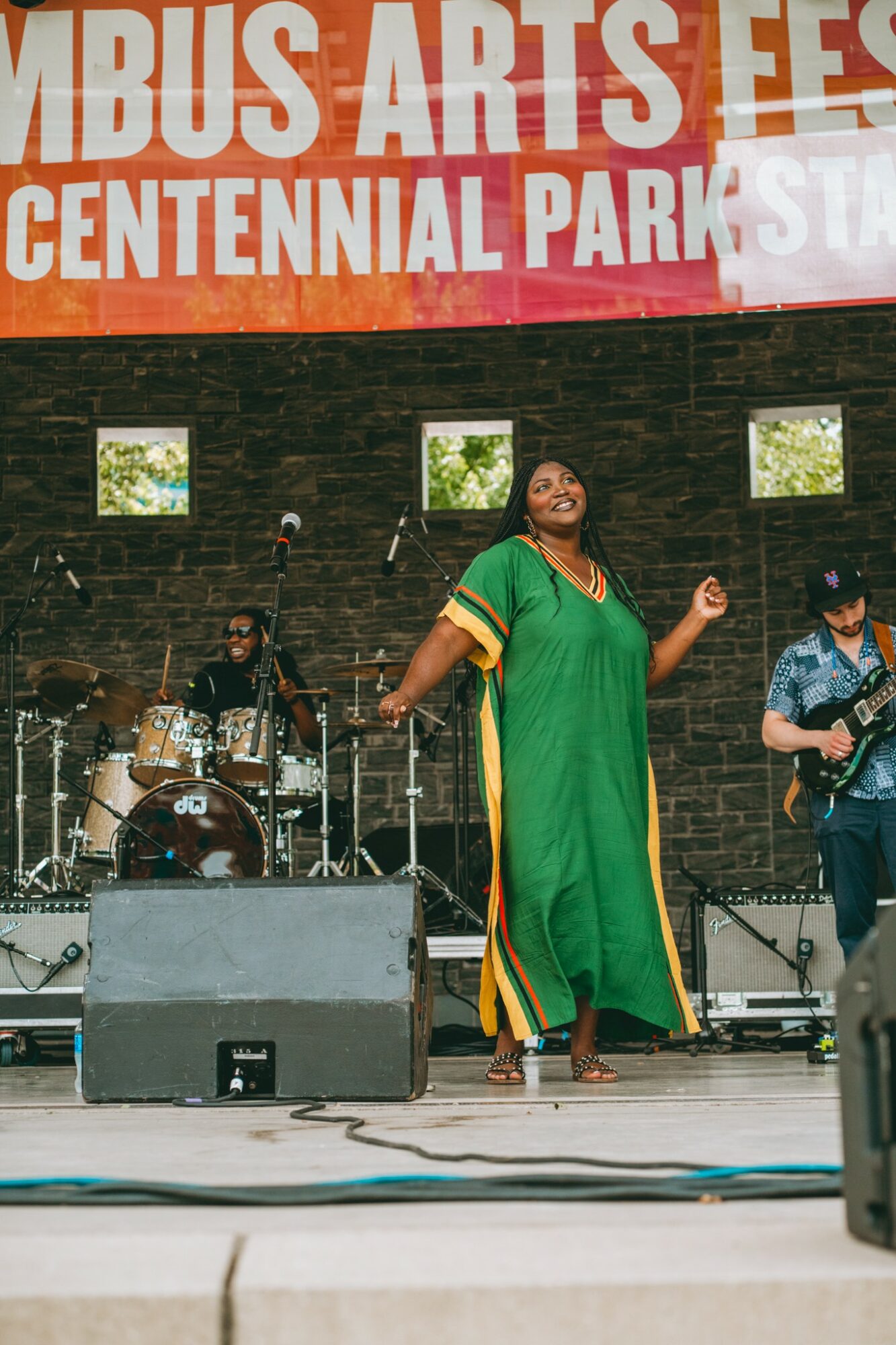 A woman in a green dress performs on stage with a band, with a brick wall and windows behind her.