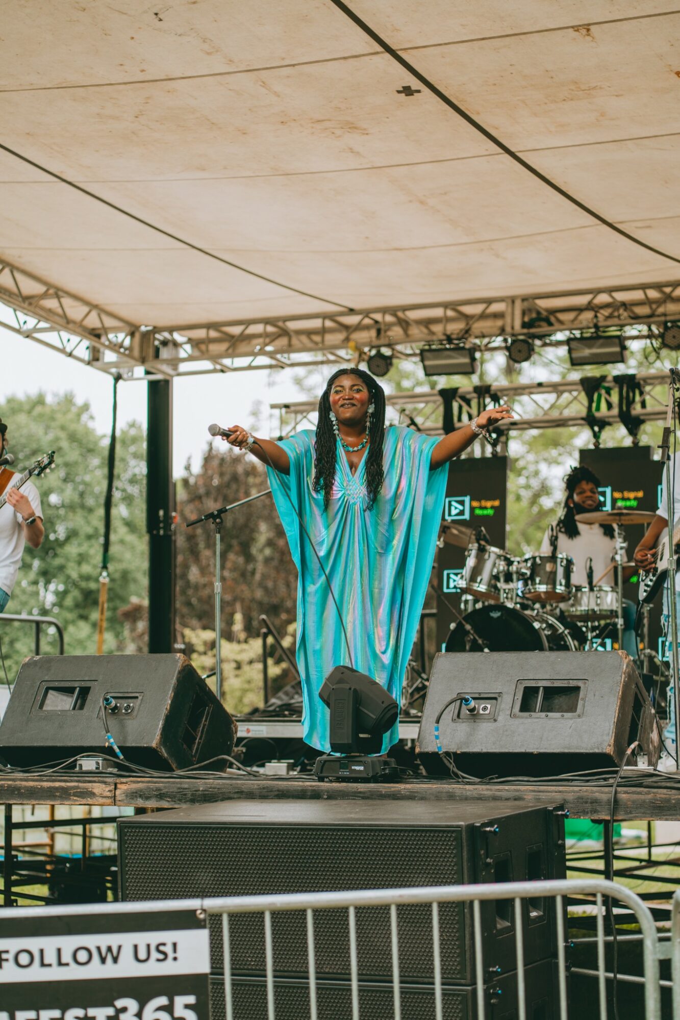Performer on stage with arms outstretched, wearing a blue dress, under a canopy, with band and equipment behind, outdoors.
