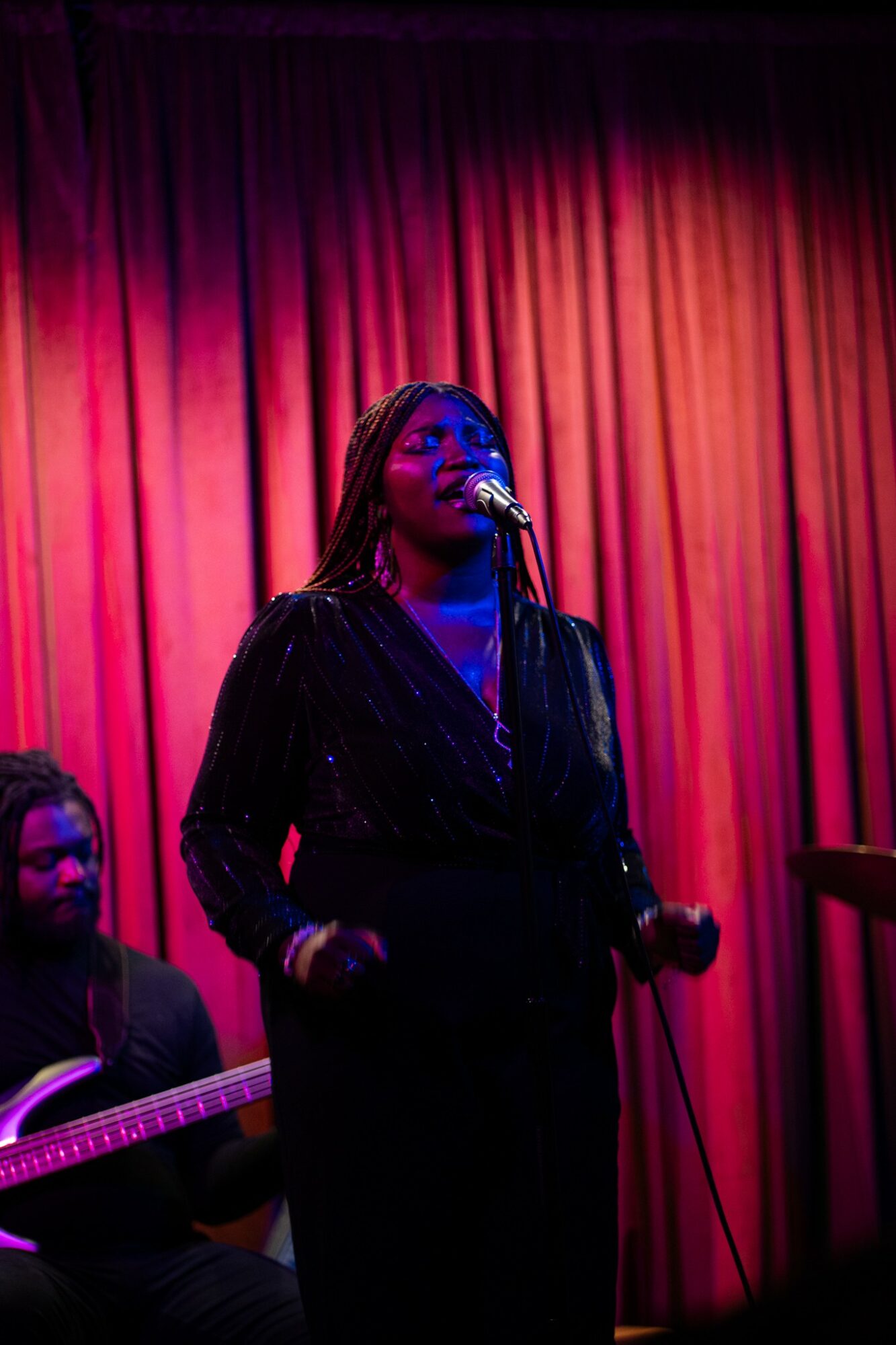 A woman singing into a microphone on stage with red curtains behind her.