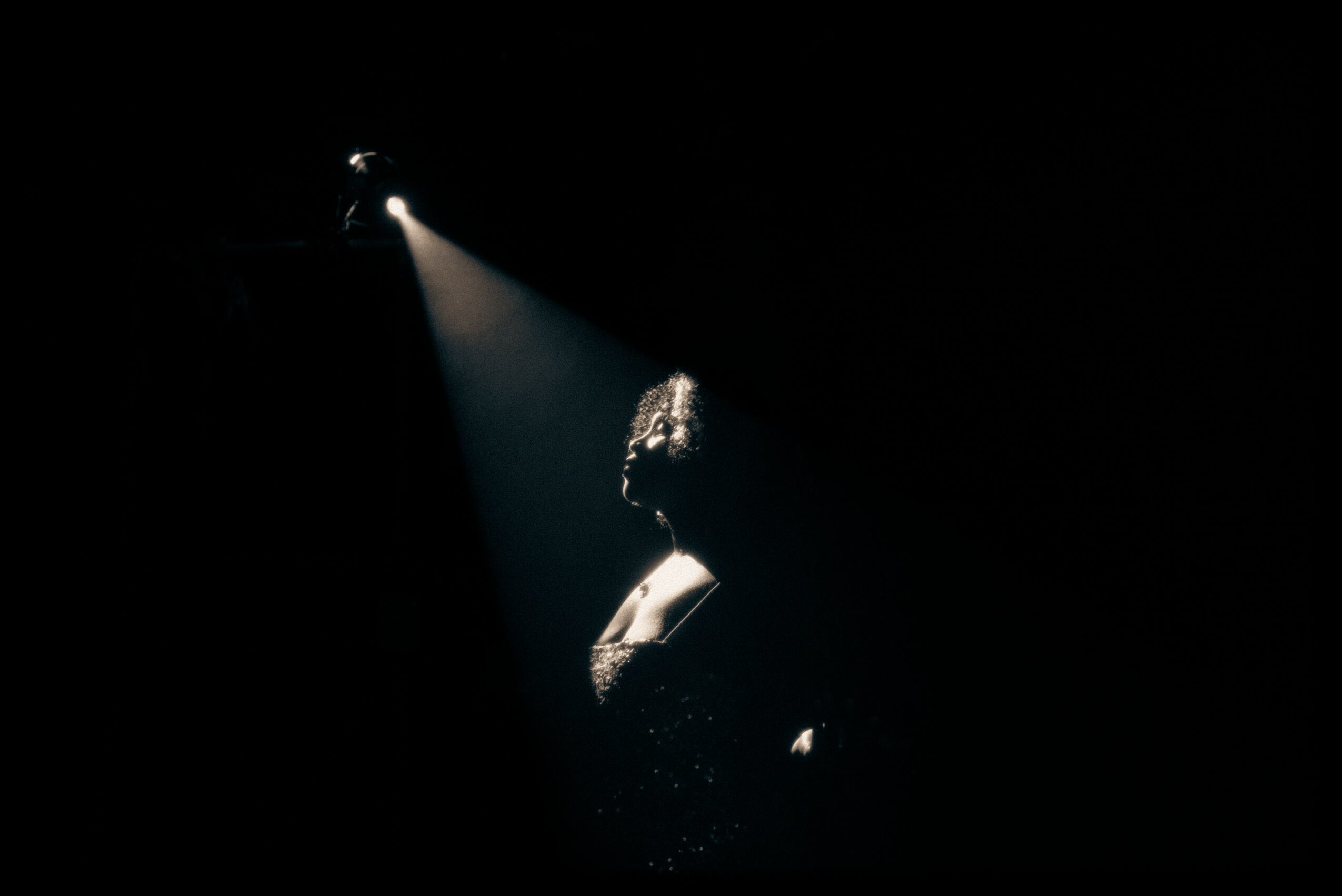 Person standing in a spotlight with dark background, face and upper body illuminated, head slightly bowed.