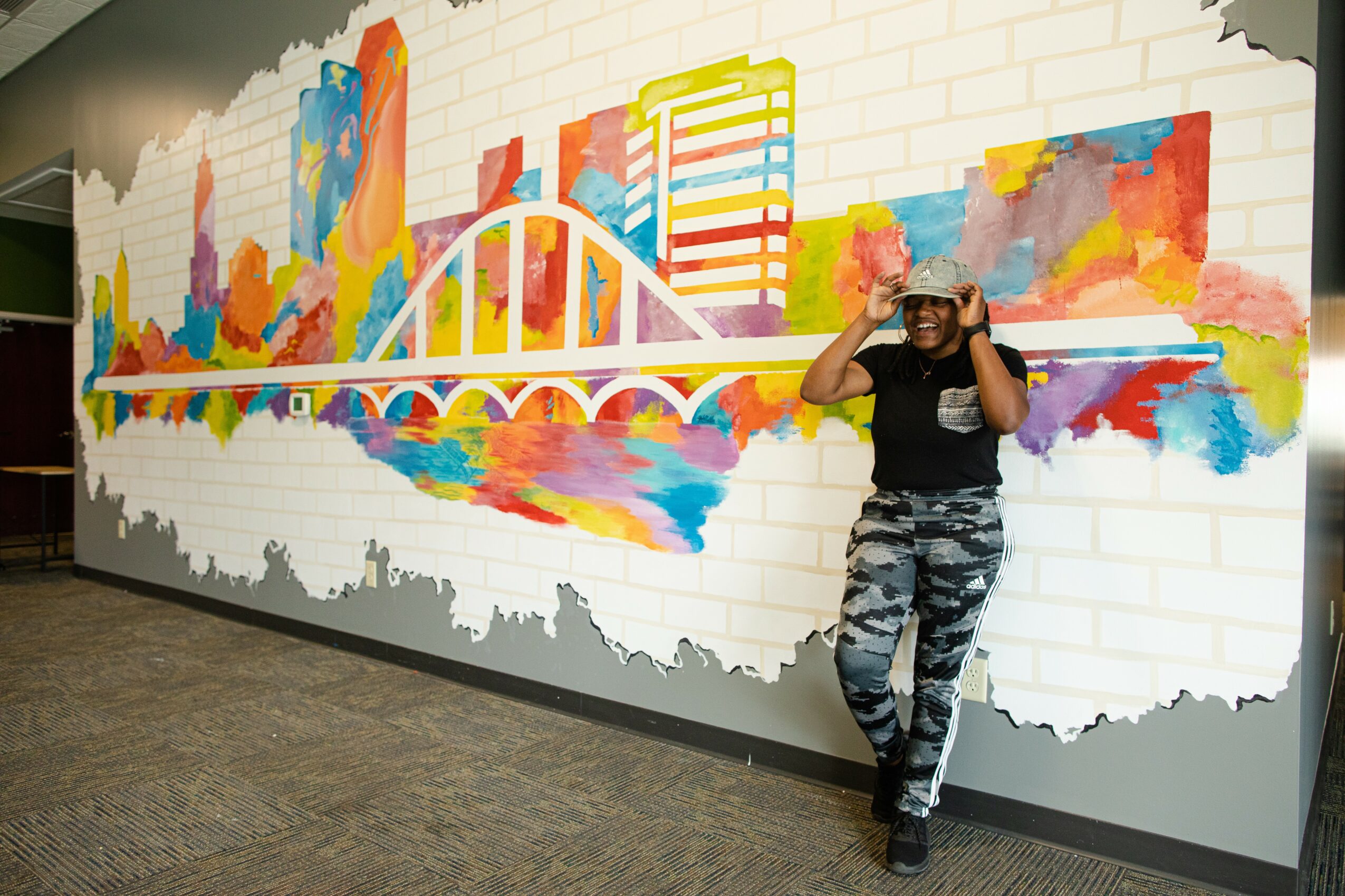 Person standing next to a colorful cityscape mural on a white brick wall, smiling and adjusting sunglasses.