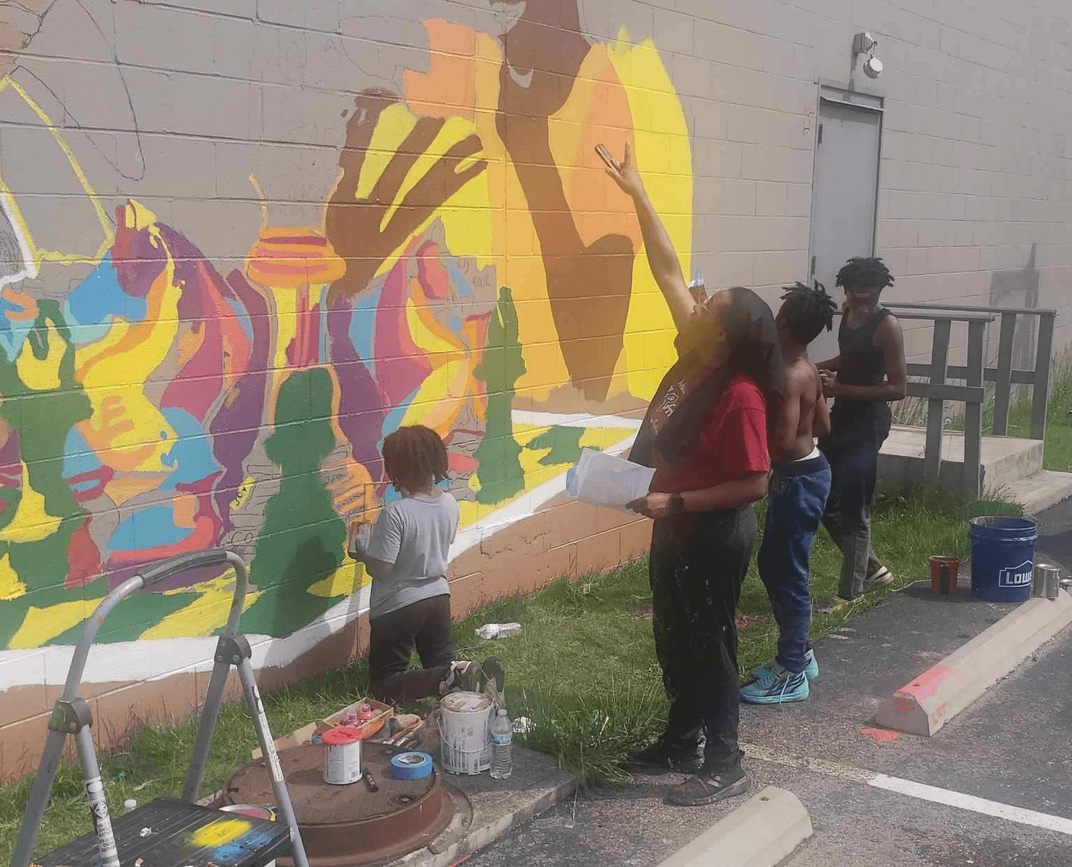 Group of people painting a colorful mural on a wall outdoors, with painting supplies nearby.