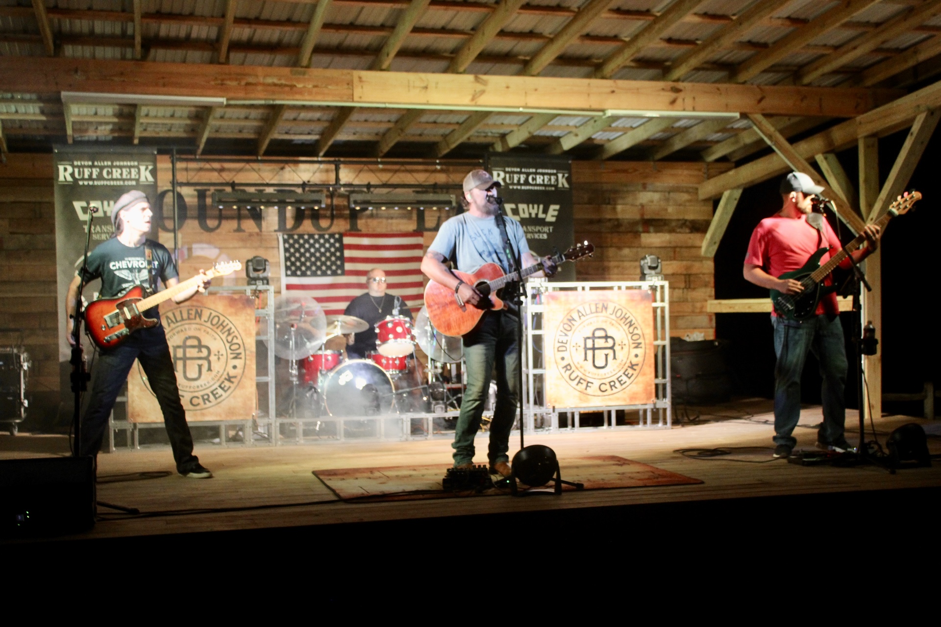Four musicians perform on stage with guitars and drums, American flag backdrop, wooden structure, and stage lighting.