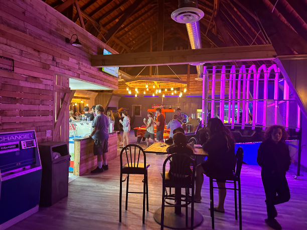 People gather in a decorated indoor space with colorful lighting, wooden walls, and seating areas, possibly a party or event venue.