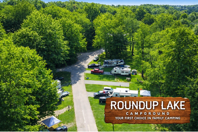 A campground with parked RVs and cars surrounded by green trees, and a dirt road leading through the area. A sign reads 'Roundup Lake Campground' with additional text.