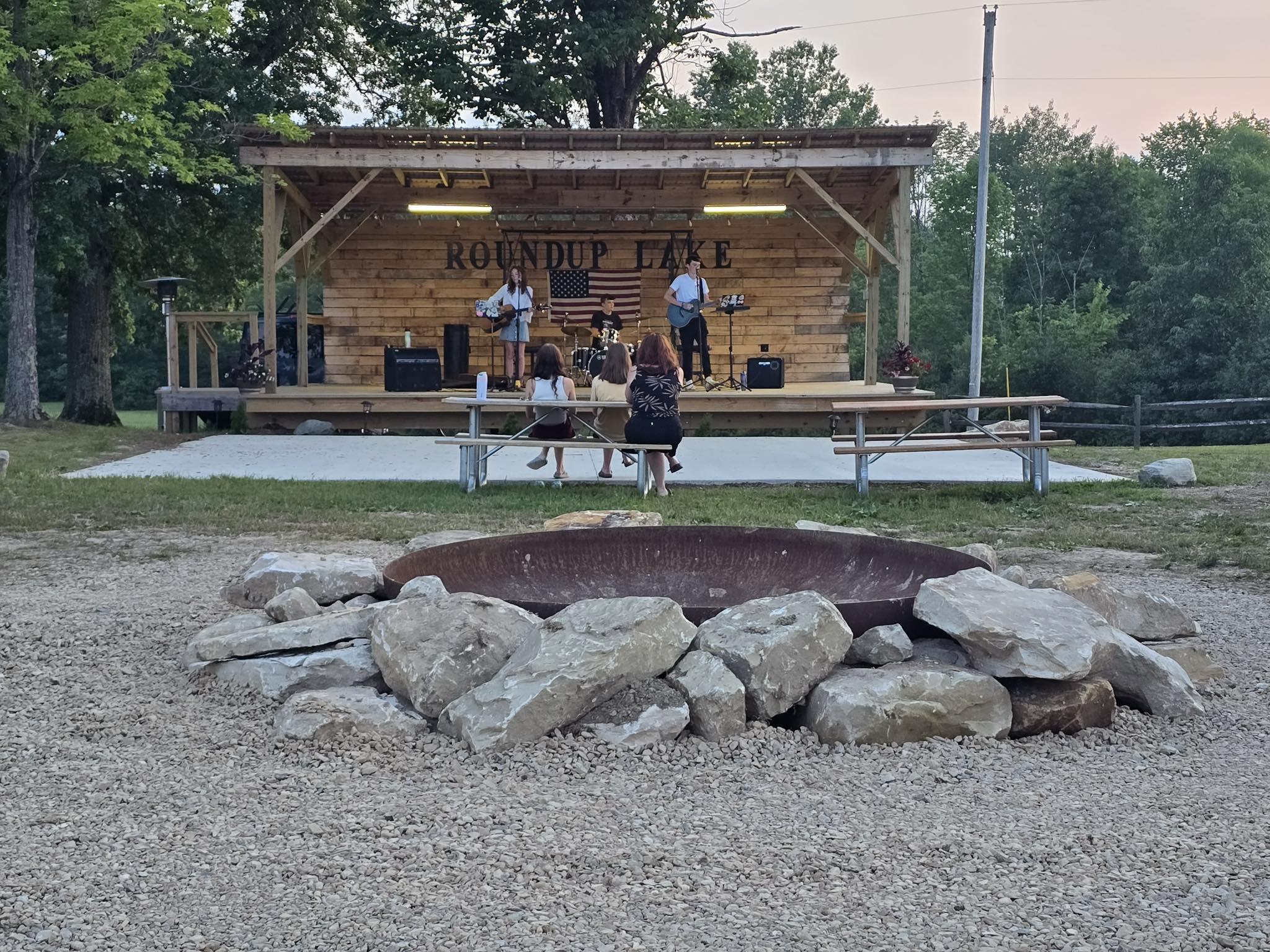 People on a wooden stage with a sign reading 'Roundup Lake,' surrounded by trees, with a fire pit and rocks in foreground.
