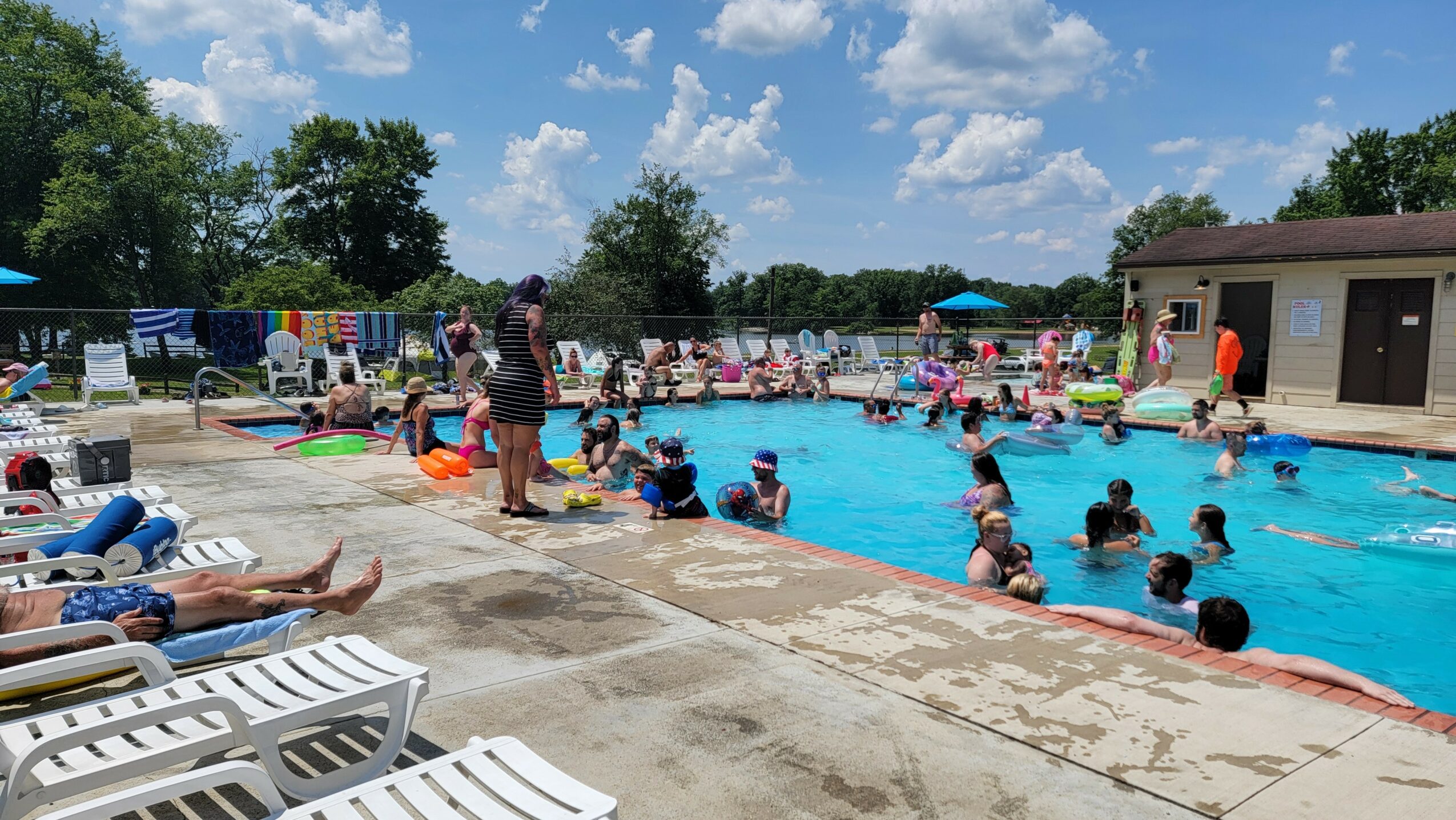 People swimming and relaxing at an outdoor pool on a sunny day, with lounge chairs and a building nearby.