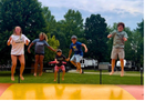 Five children jumping over a colorful obstacle outdoors with trees and a cloudy sky in the background.