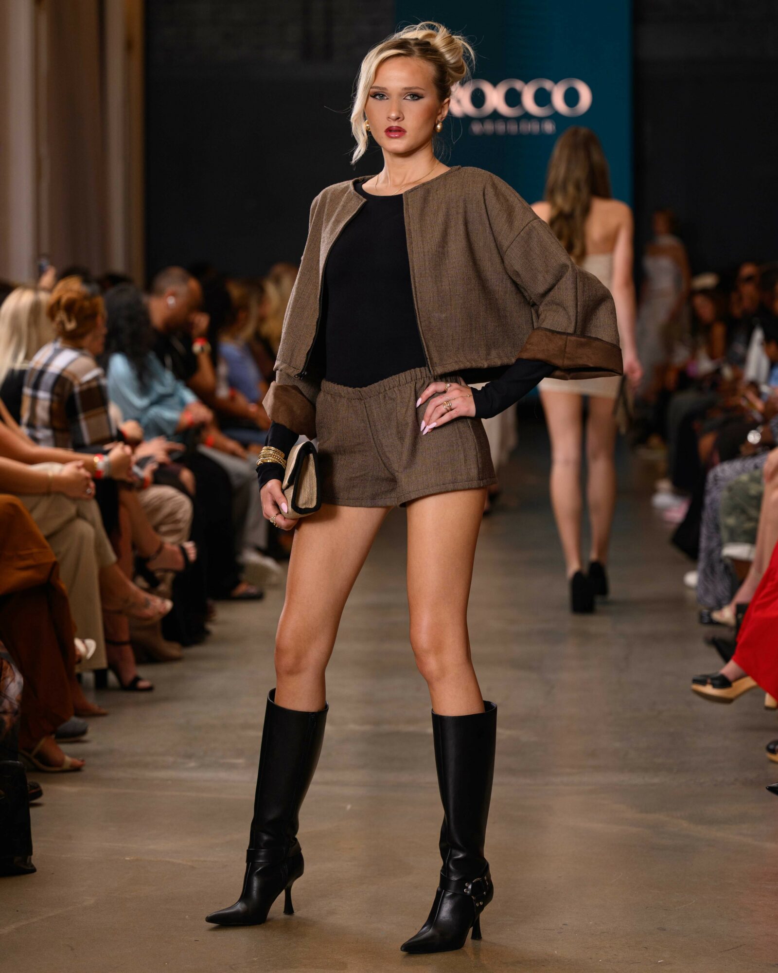 Fashion model walking runway in black top, brown cape, shorts, and knee-high black boots, audience seated on sides, runway background.
