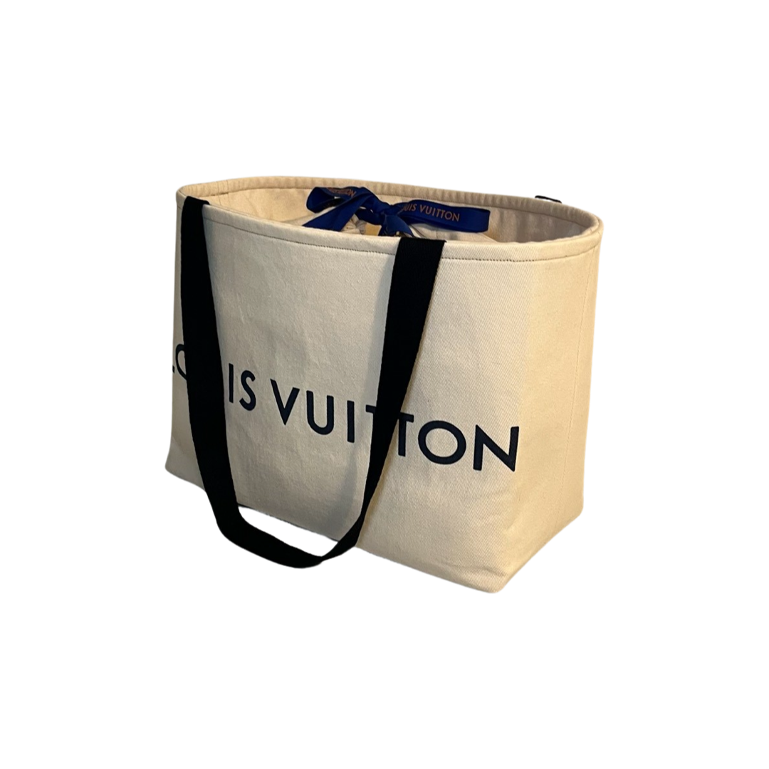Cream-colored tote bag with black handles and blue ribbon, partially visible text, on a white background.