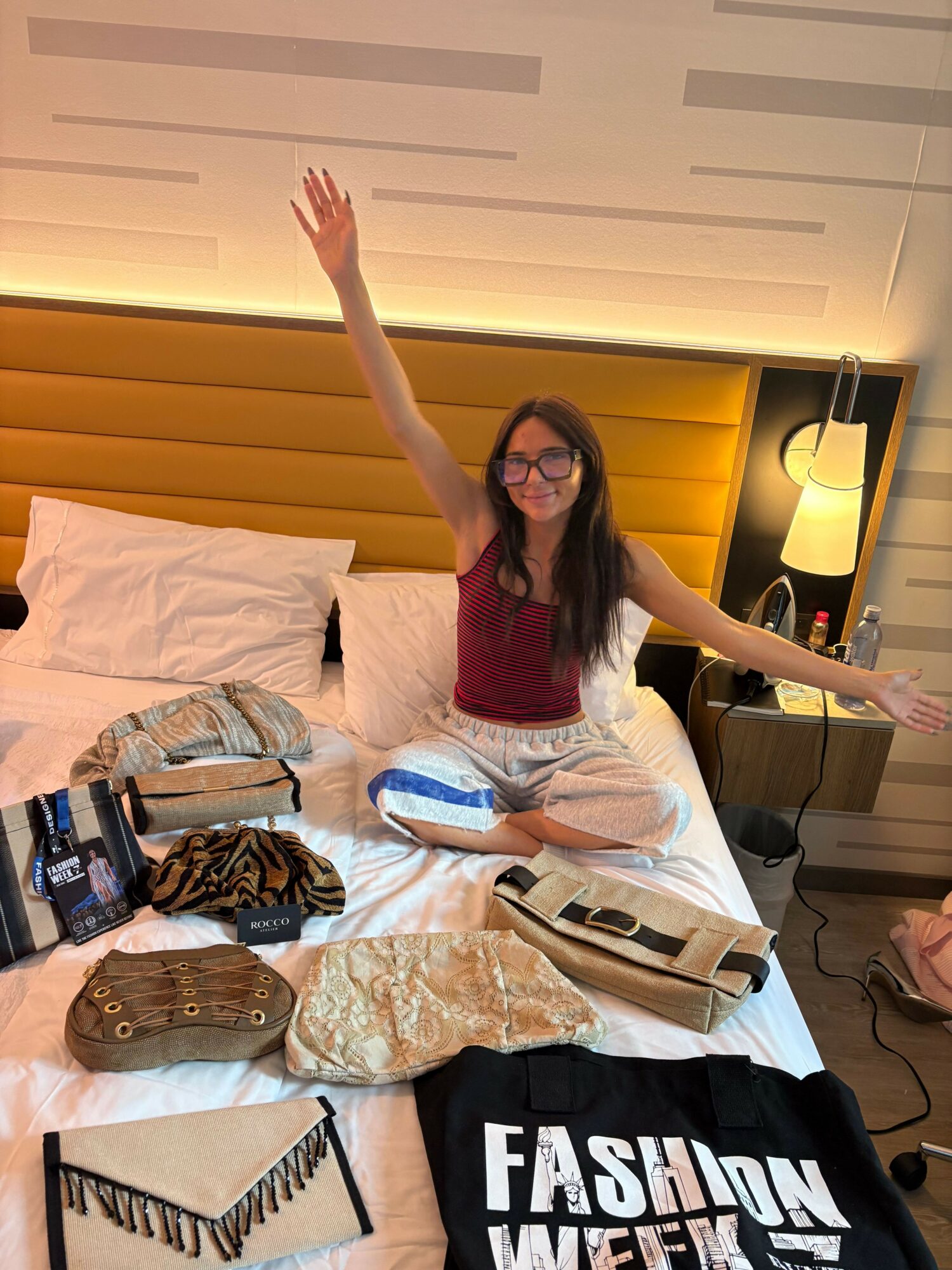 Person with long dark hair and glasses, wearing a red top, sitting on a bed with arms raised, surrounded by bags and accessories.