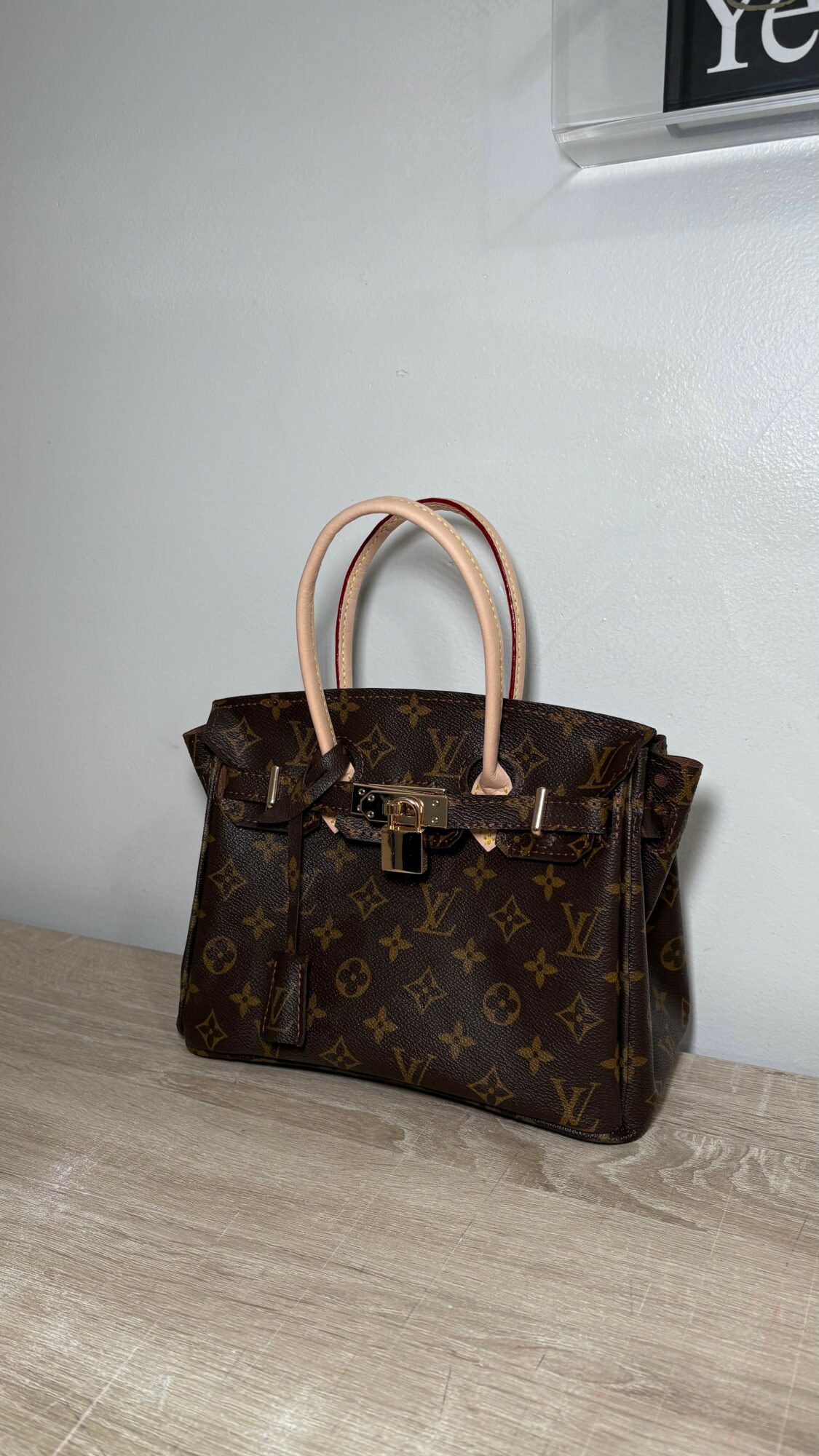 Louis Vuitton handbag with monogram pattern and light-colored handles on a wooden surface, against a plain wall.