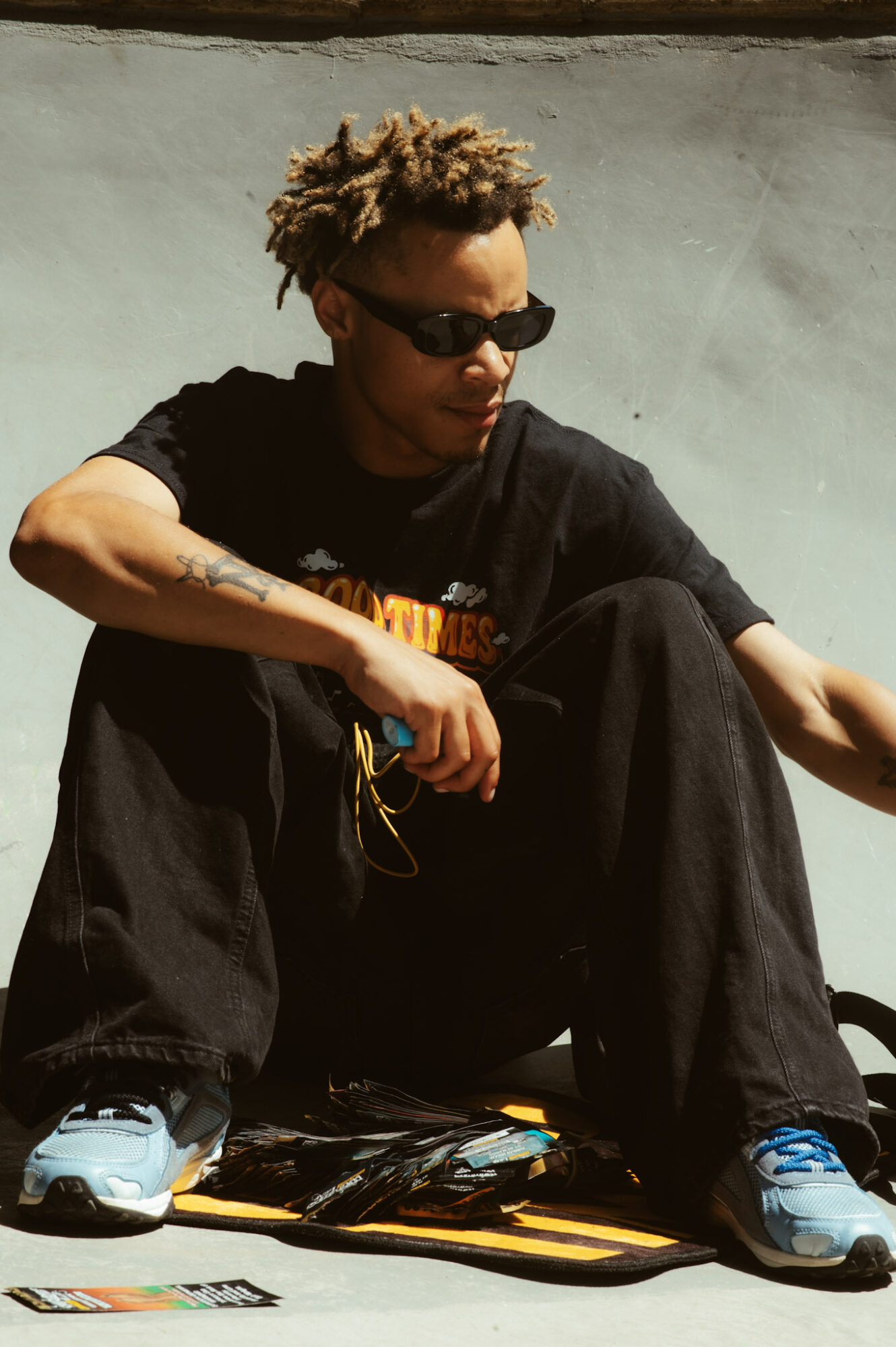 Person with curly hair wearing sunglasses, black T-shirt, and black pants, sitting outdoors on a skateboard, with a plain wall background.
