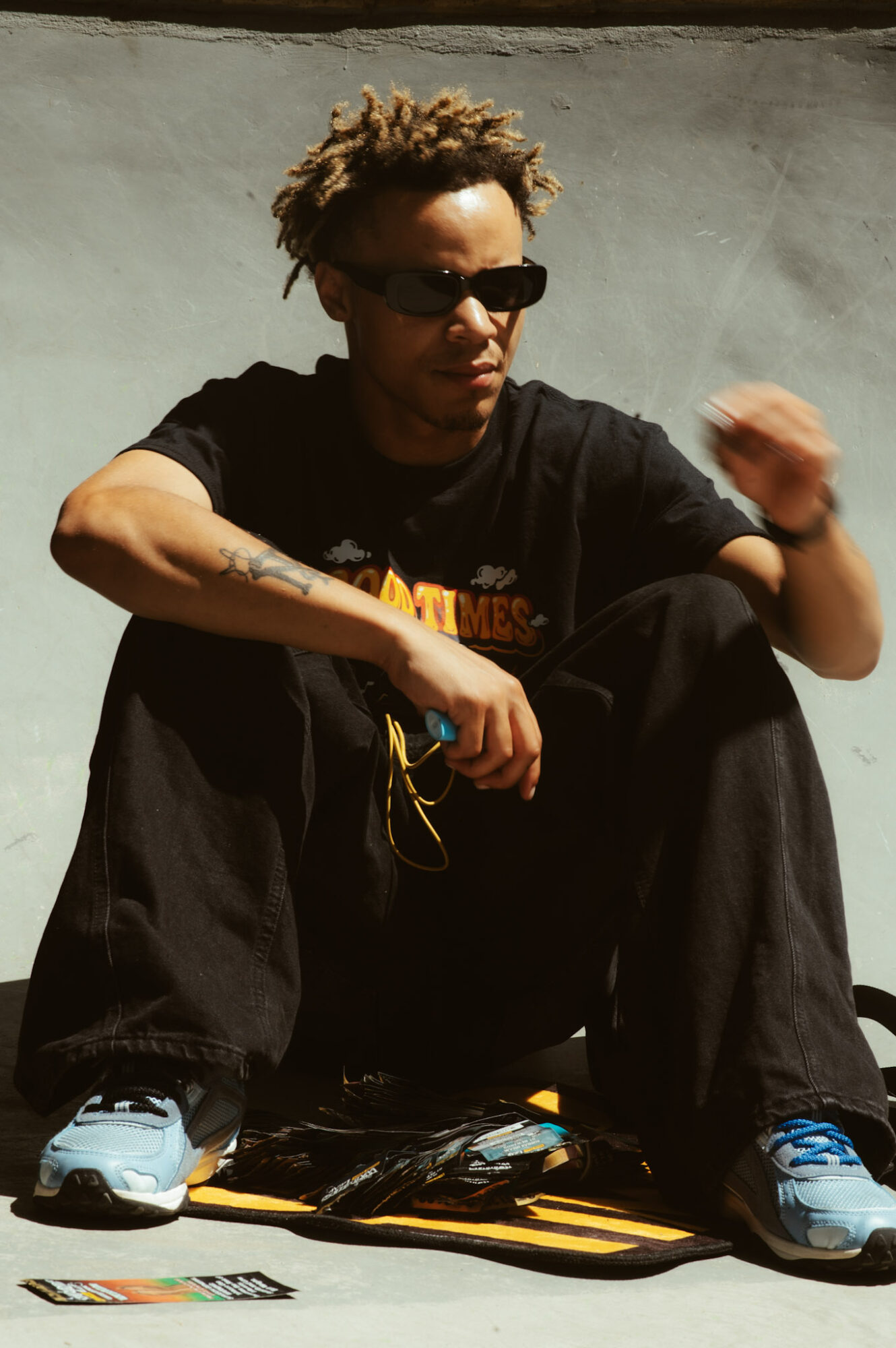 Person with curly hair wearing sunglasses, black t-shirt, and black pants, sitting on the ground with a gray background.