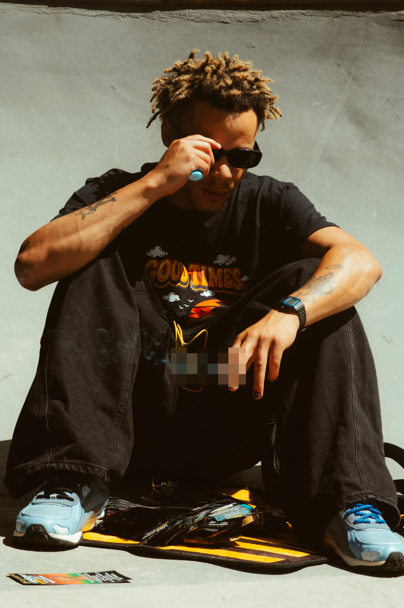 Person with sunglasses squatting, wearing a black T-shirt, black pants, and sneakers, against a plain background.