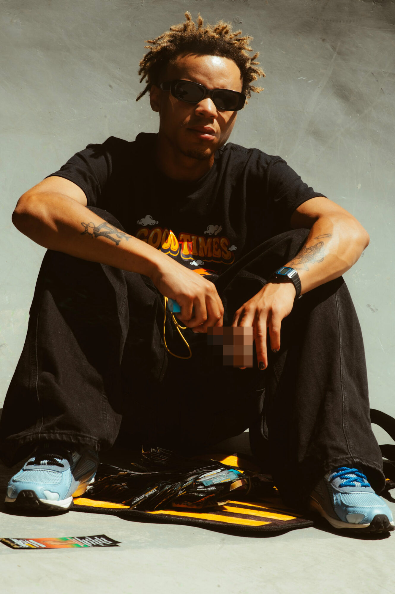 Person with sunglasses sitting on a skateboard, holding a phone, against a plain background.