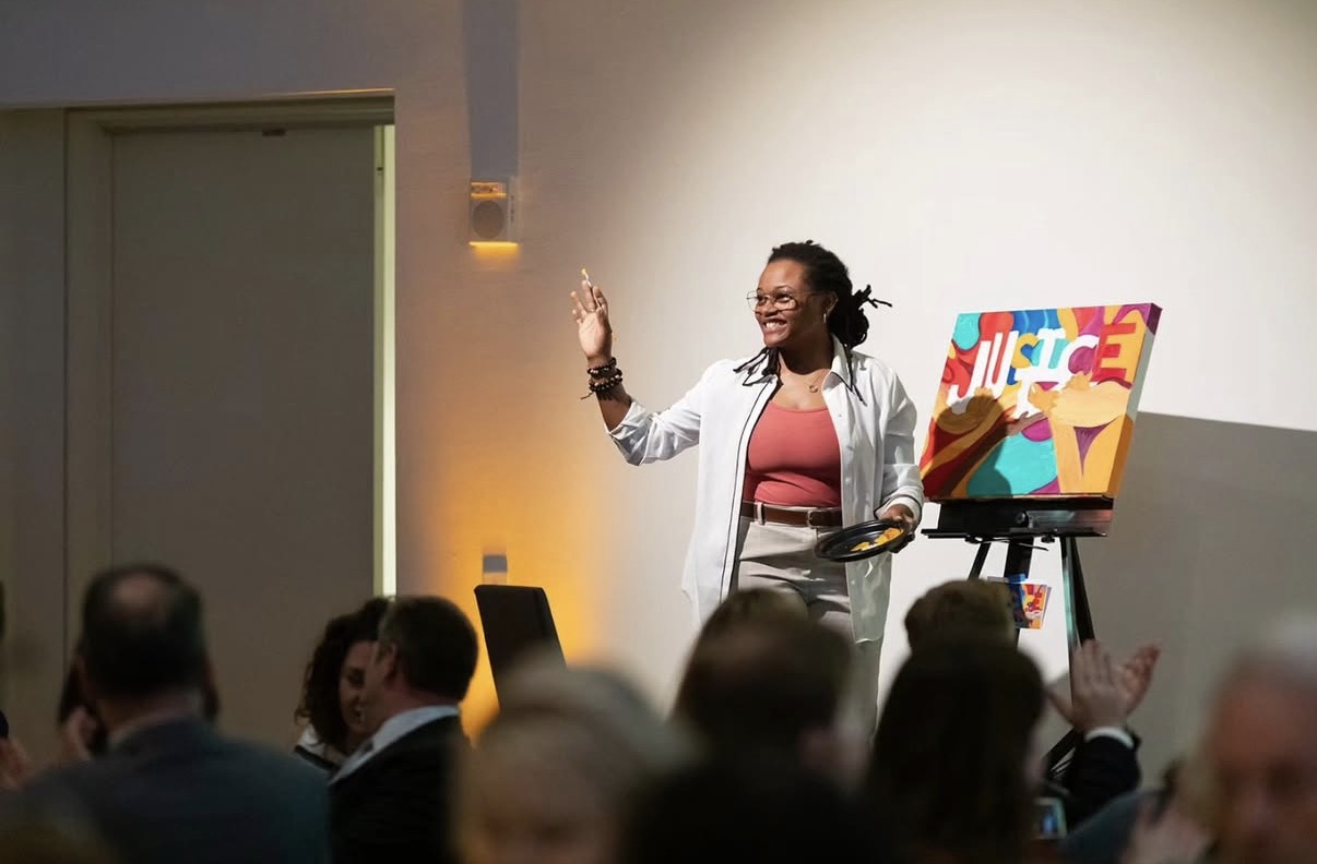 Woman giving a presentation on stage with a colorful artwork on an easel, audience seated in front.