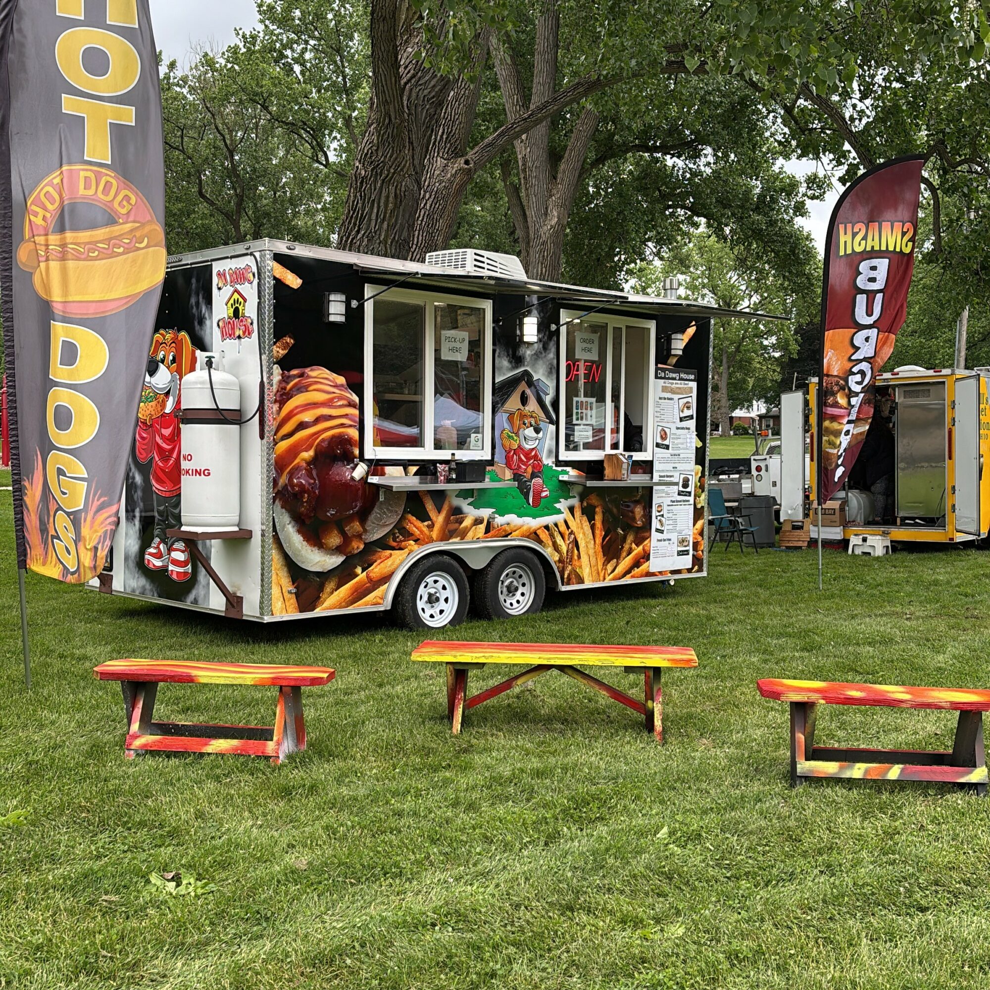 Food truck with colorful graphics, two benches in front, trees and flags in the background, grassy area.