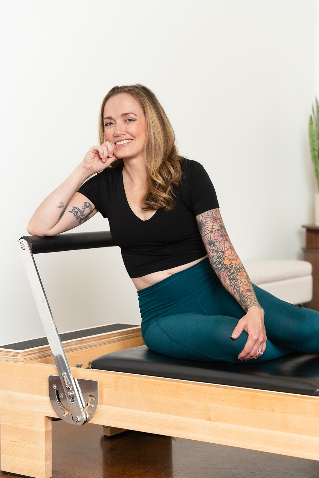 Woman with tattoos sitting on a Pilates reformer machine, smiling, in a bright room with a plant in the background.