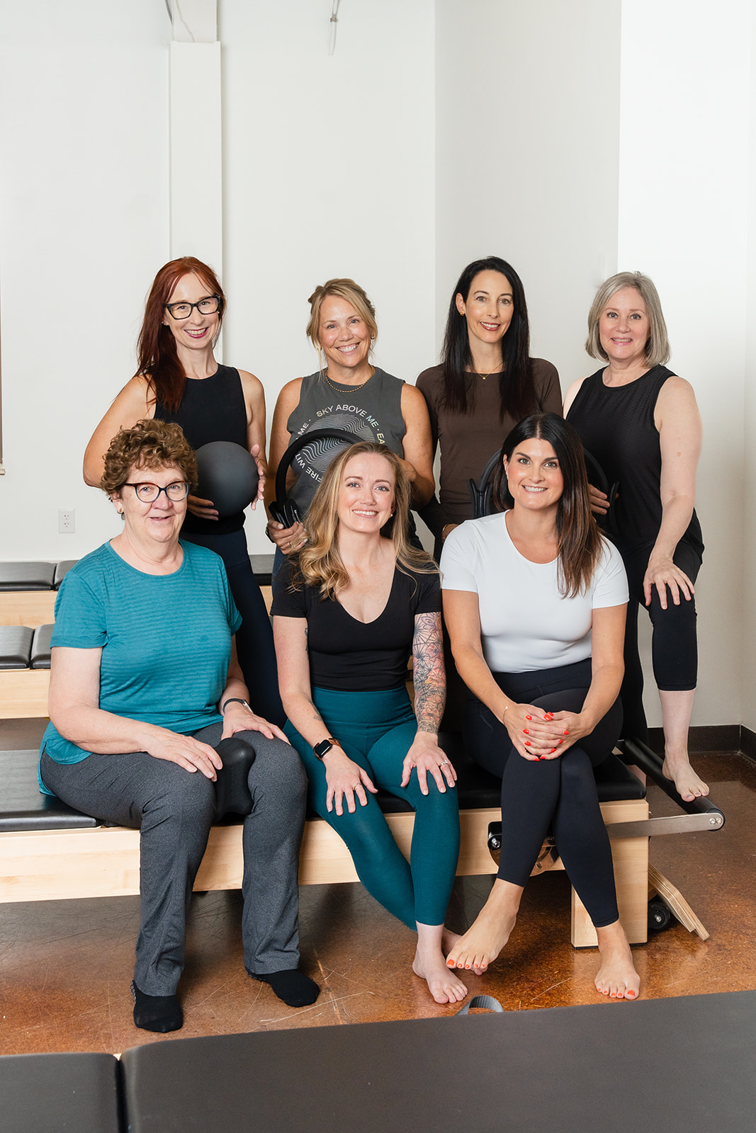 Seven women posing together in a room with white walls, some sitting and some standing, smiling at the camera.