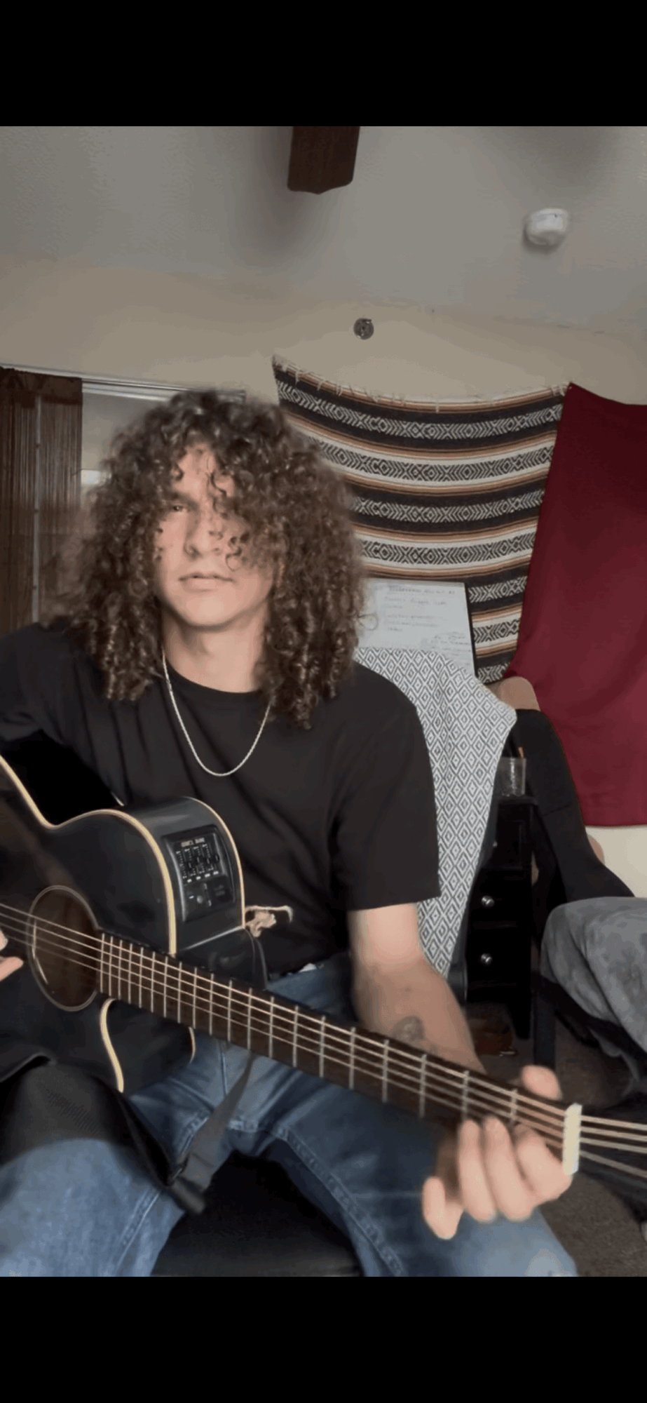 Person with curly hair playing an acoustic guitar indoors, sitting on a chair, with a striped curtain and red fabric in the background.