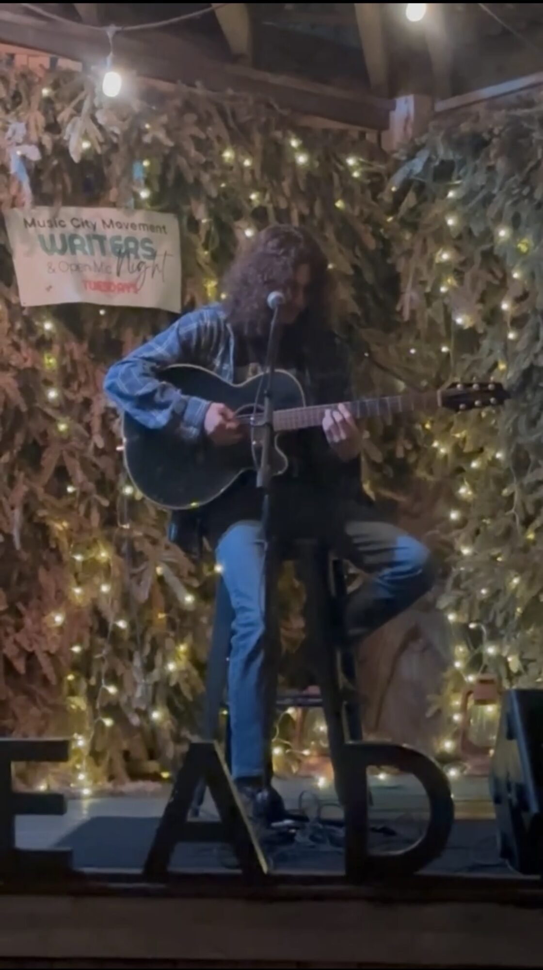 Person playing guitar on stage with string lights and a sign in the background, surrounded by decorative elements.