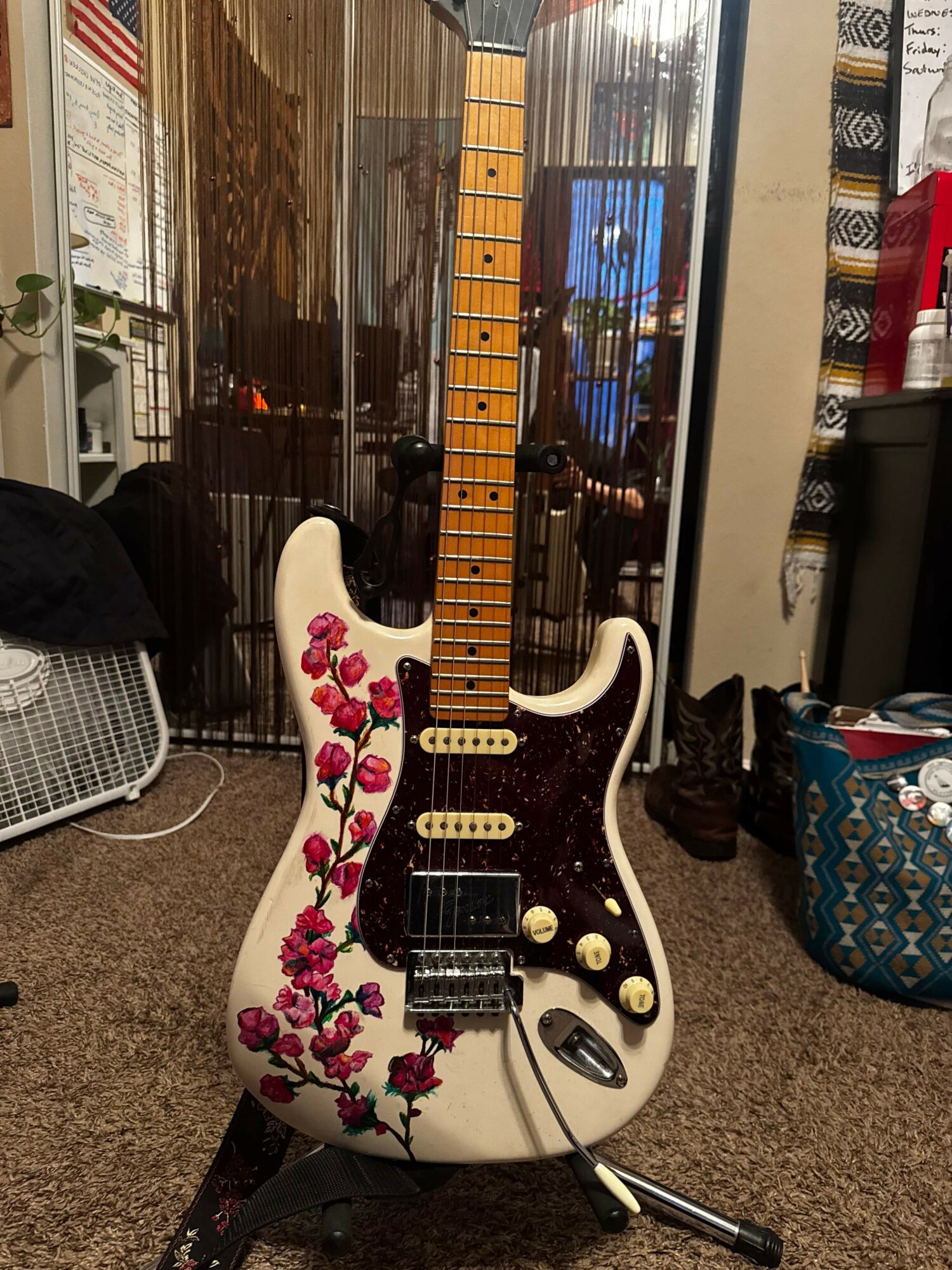 Electric guitar with floral design, white body, and wooden neck, standing on a carpeted floor in a room.