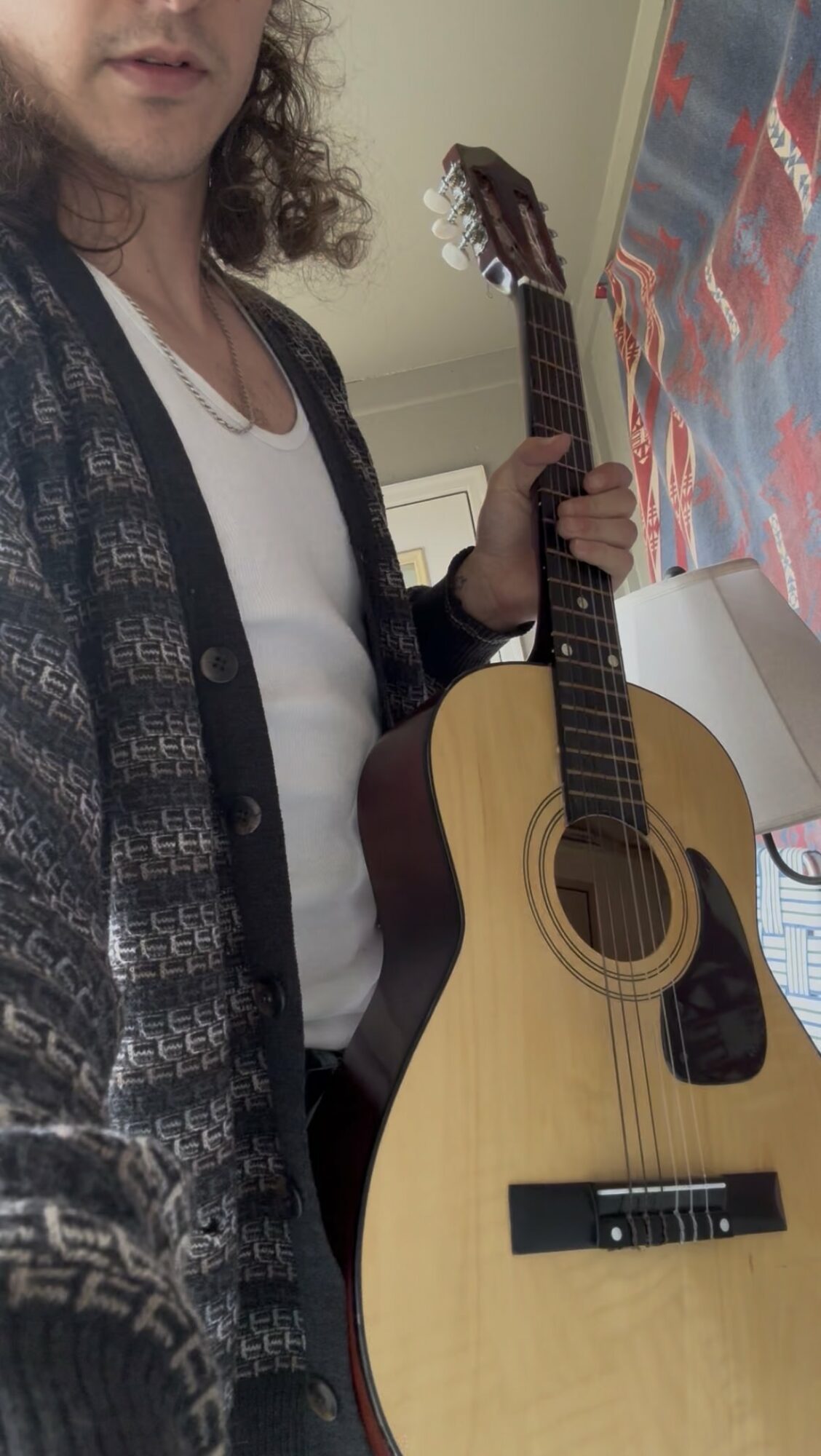 Person holding an acoustic guitar, wearing a patterned cardigan and white shirt, standing indoors near a colorful wall.
