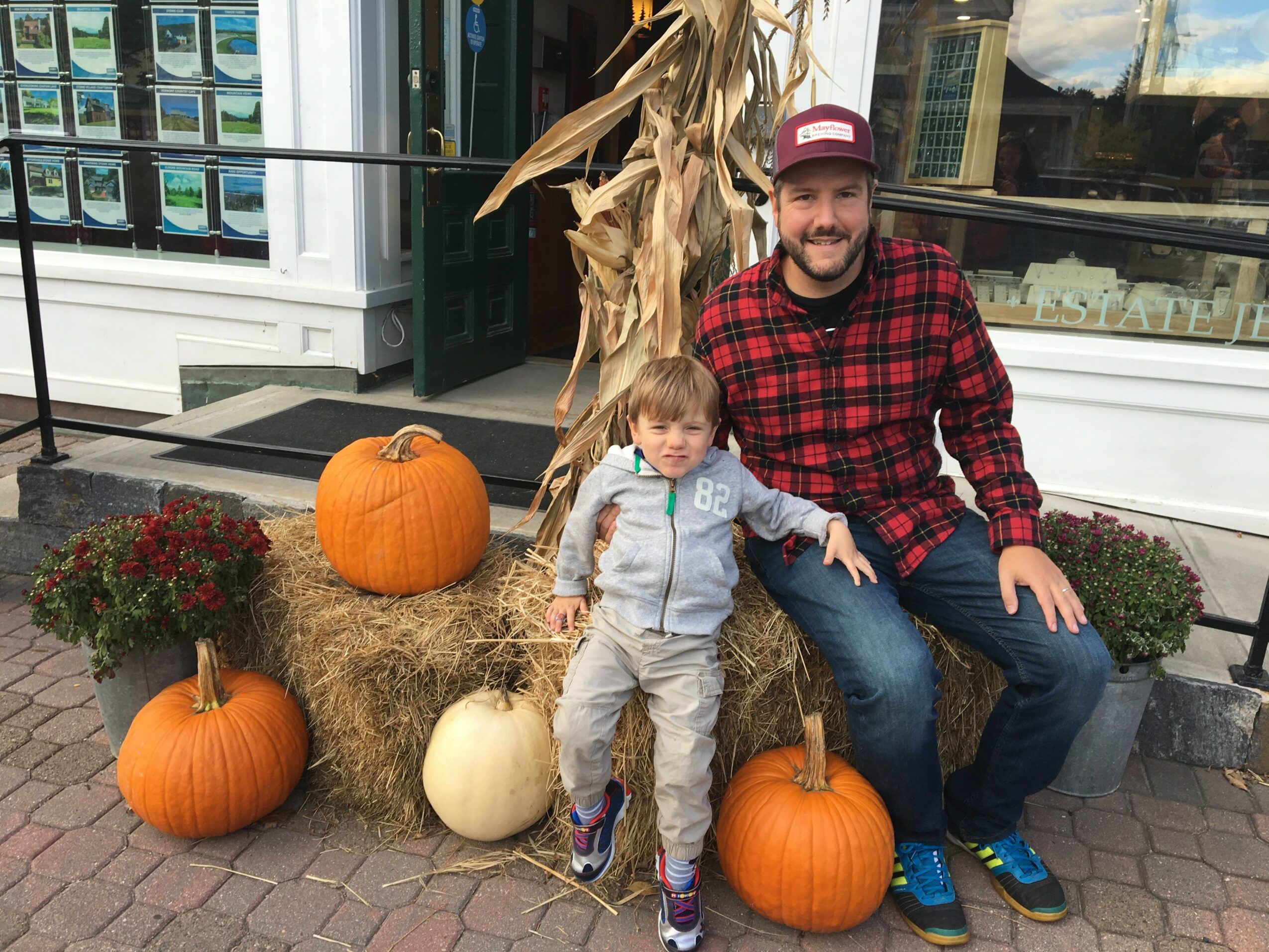 A man and a child sitting on hay bales surrounded by pumpkins and fall decorations outside a store.