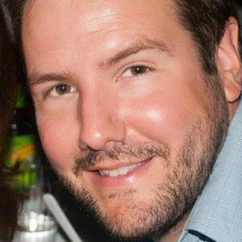 Close-up of a smiling man with short dark hair and facial hair, wearing a light blue shirt.
