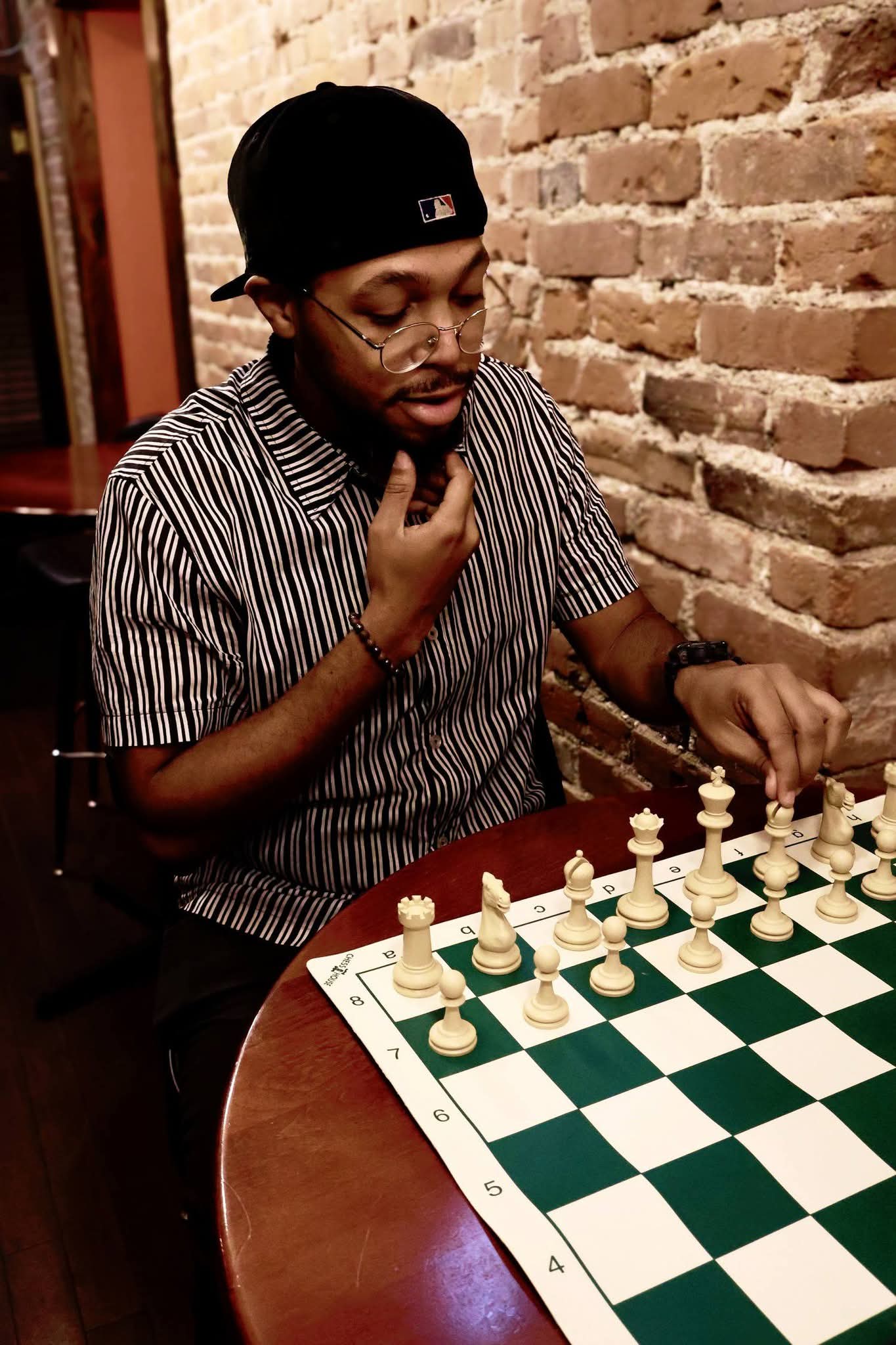 Person with glasses and a black cap playing chess against a brick wall background.