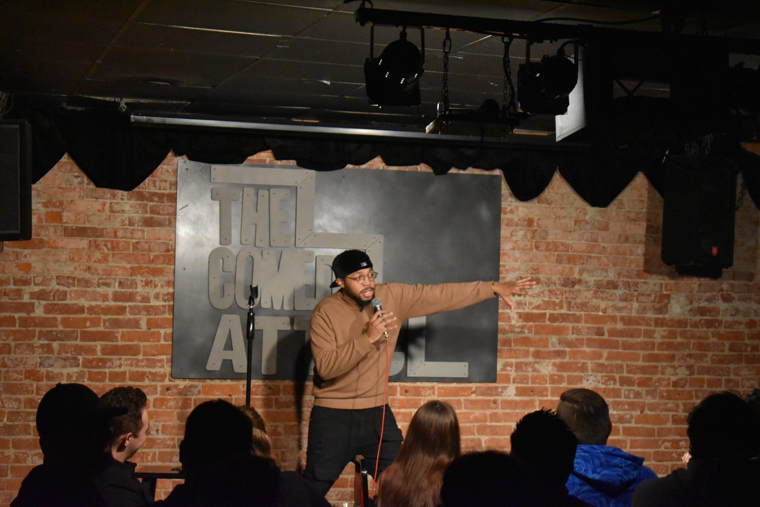 Person performing stand-up comedy on stage with brick wall background and audience watching.