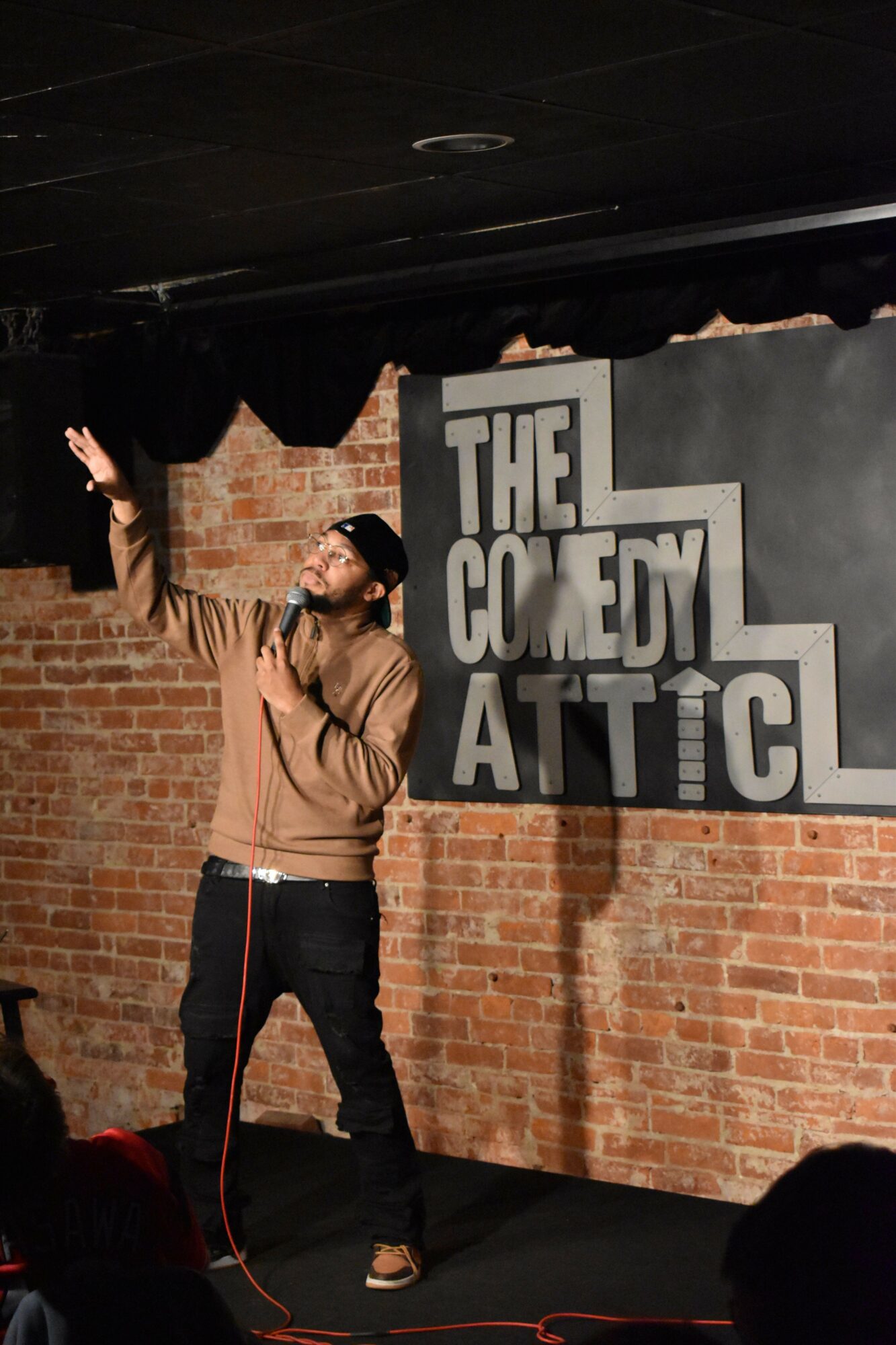 Person on stage gesturing with hand, brick wall background, sign reading 'THE COMEDY AT THE' behind them.