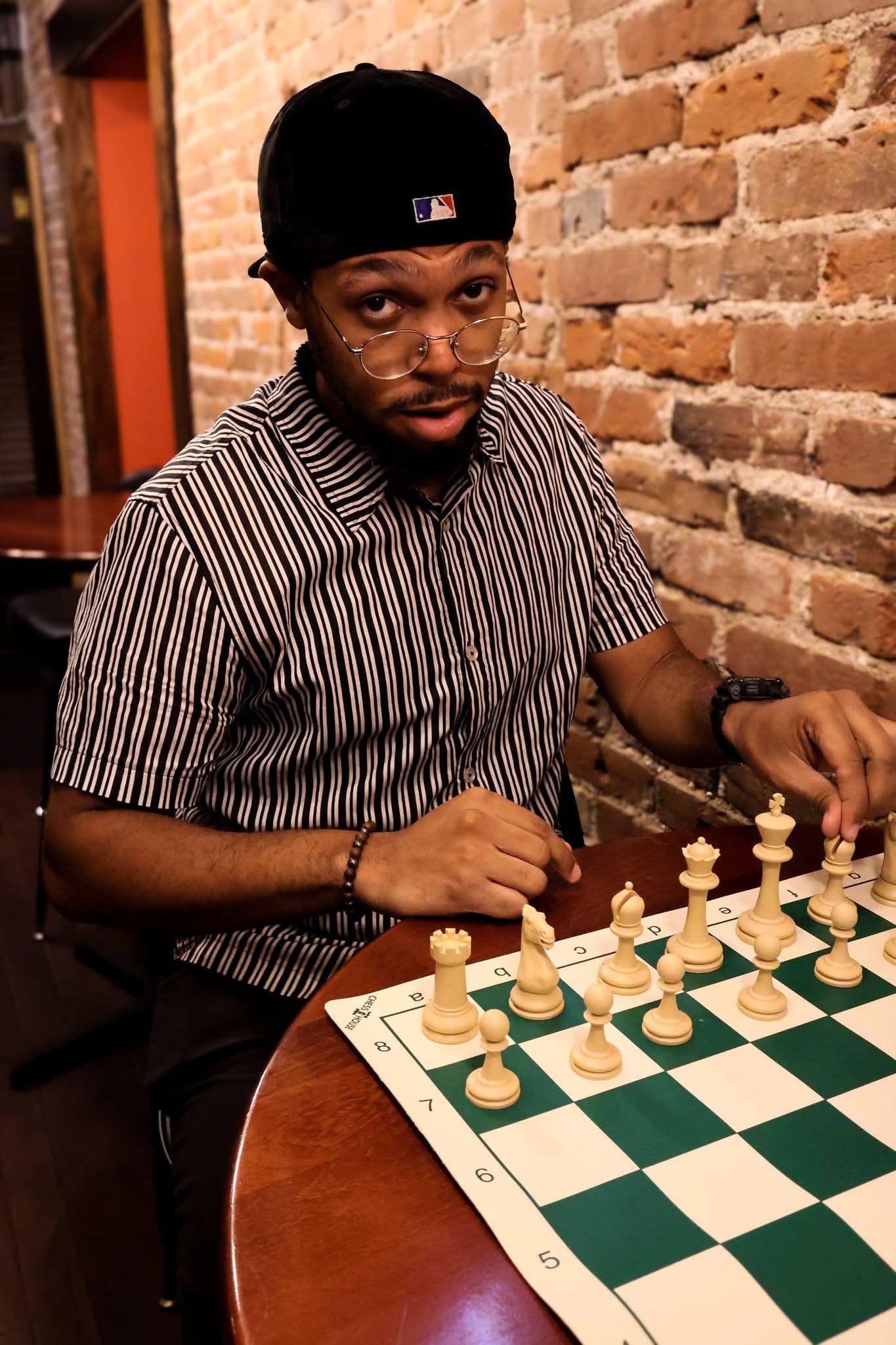 Man with a beard and glasses wearing a black cap and striped shirt, sitting at a table with a chessboard, against a brick wall.