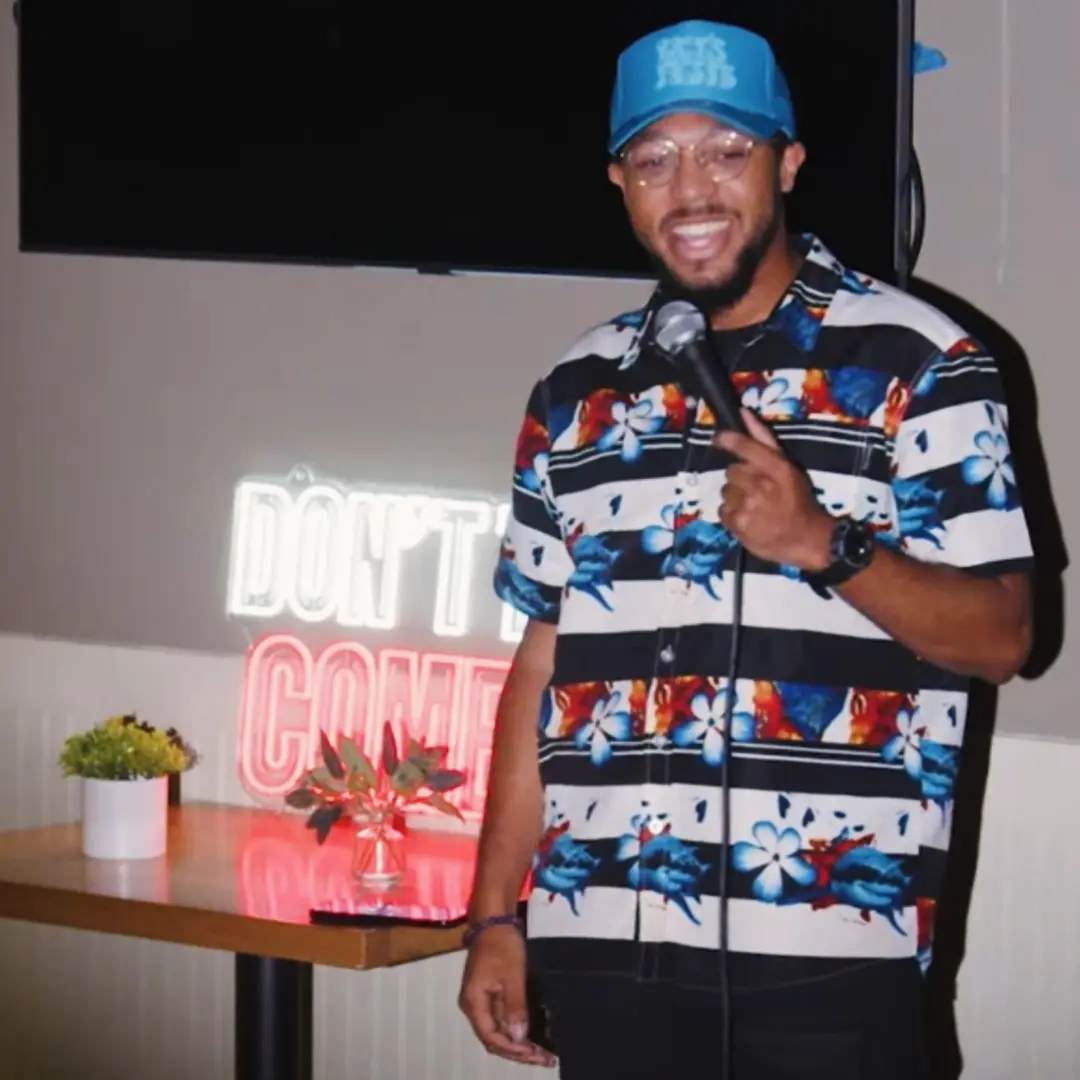 Man wearing a blue cap and floral shirt holding a microphone, smiling, standing indoors near a table with a plant and a neon sign.