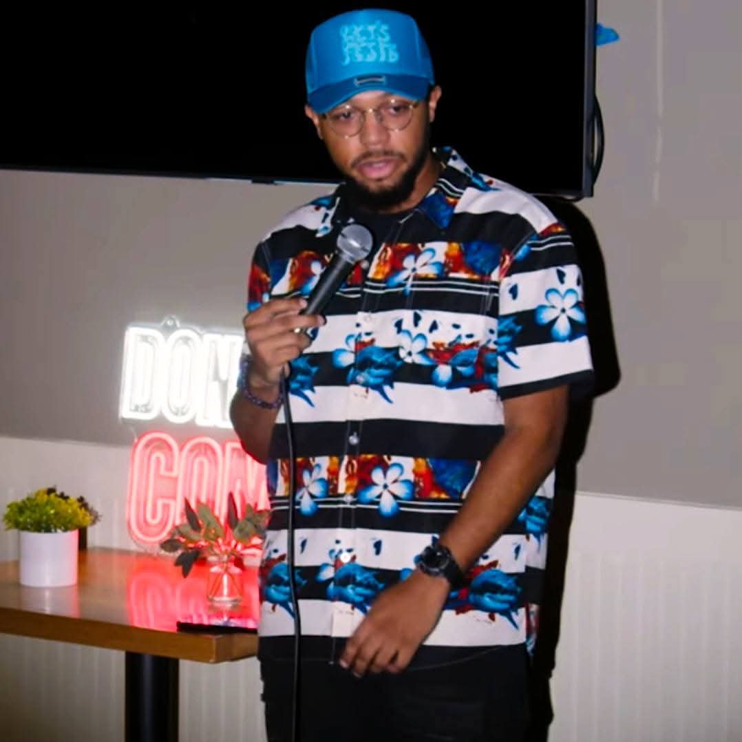 Man wearing a blue cap and floral shirt holding a microphone, standing indoors with a neon sign and table nearby.