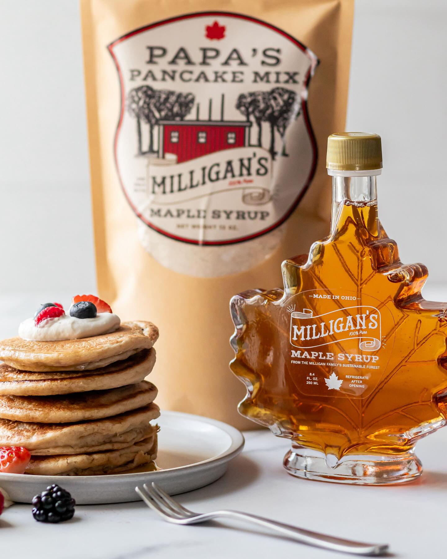Stack of pancakes with berries on a plate, maple syrup in a maple leaf-shaped bottle, and pancake mix bag in background.