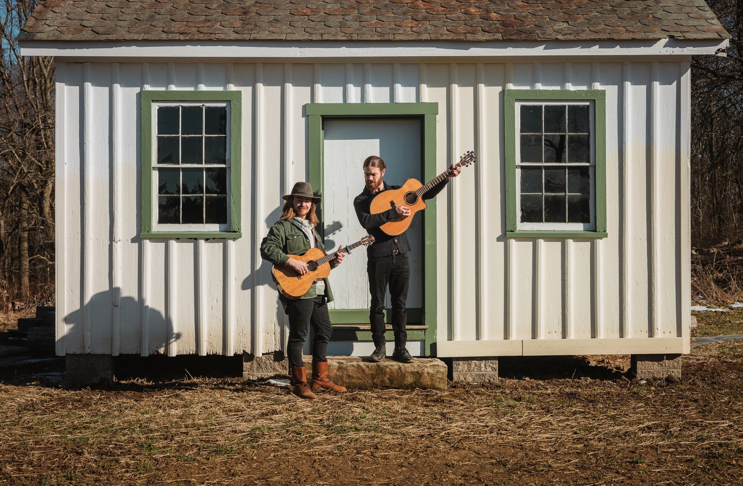 Two people playing guitars outside a small white building with green window frames and door, sunlight casting shadows.