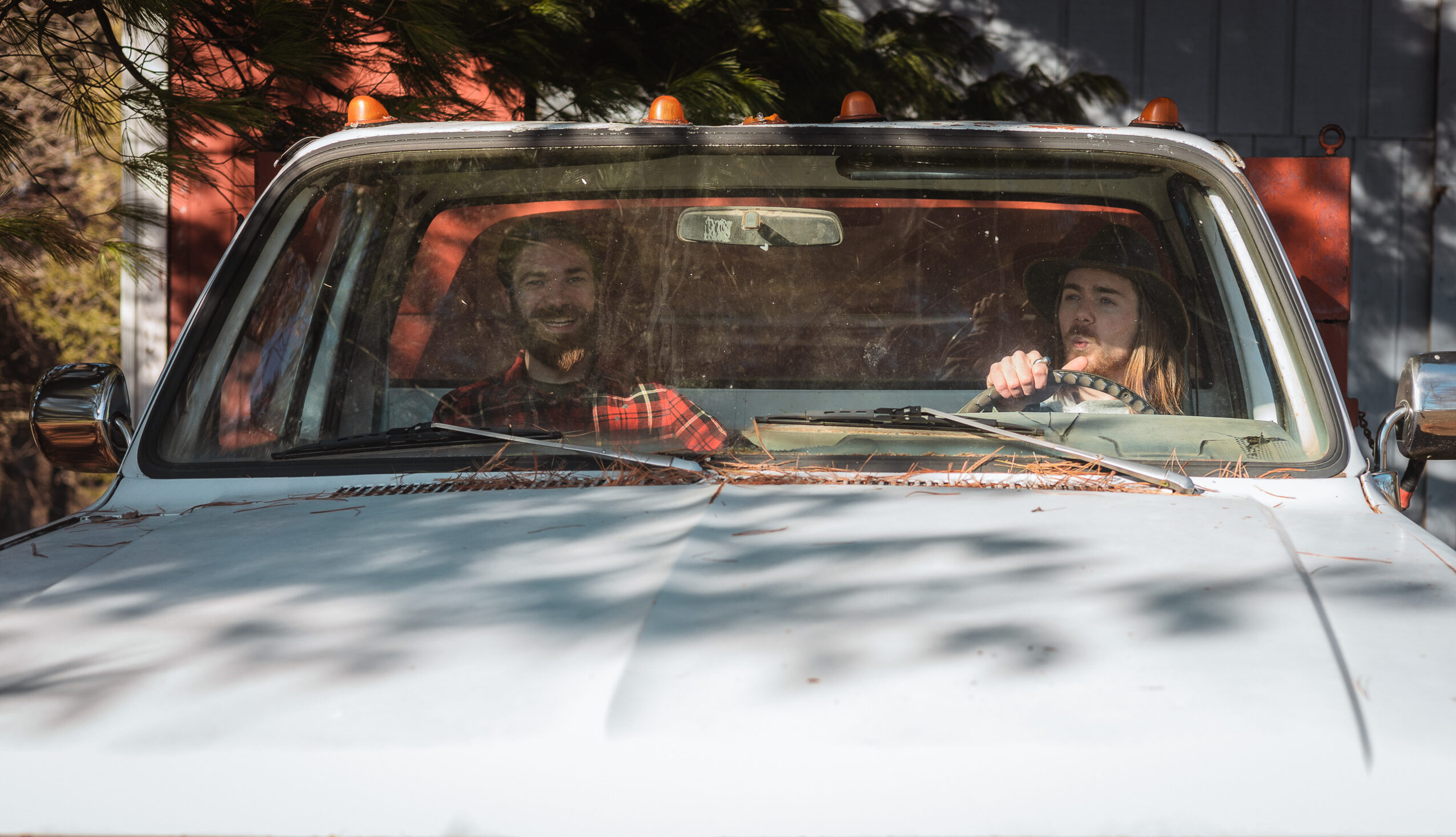 Two people sitting inside a car, visible through the windshield, with trees and sunlight outside.