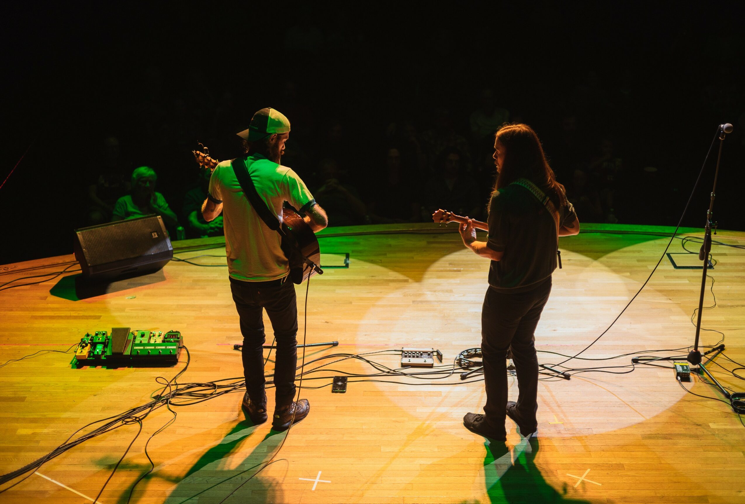 Two musicians playing guitars on stage with green lighting, cables on the floor, and a dark background.