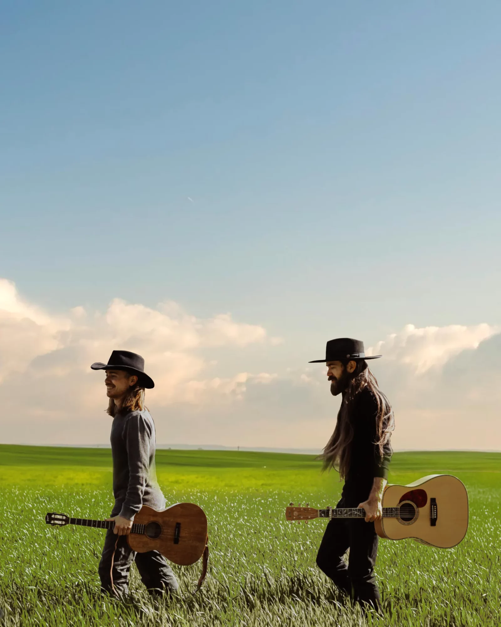 Two people walking in a grassy field holding guitars, wearing hats, with a blue sky and clouds above.