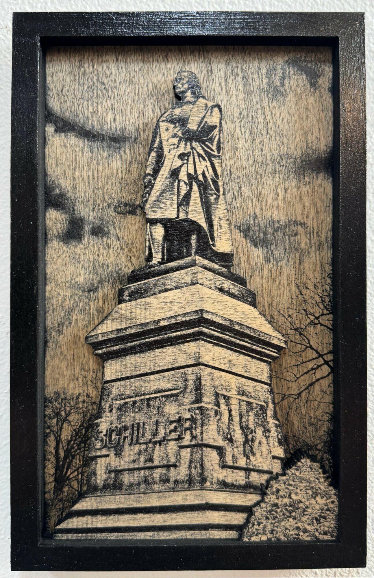 Framed photograph of a statue of a person standing on a pedestal with carved details, against a cloudy sky background.