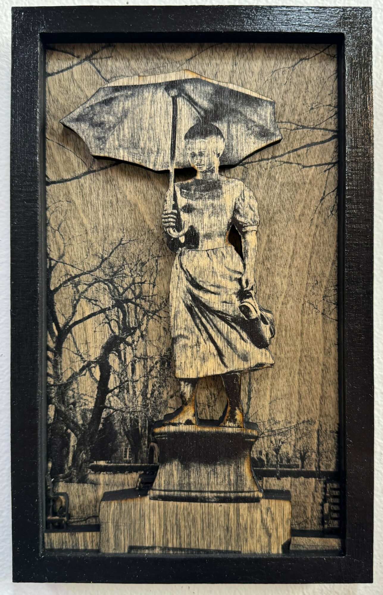 Wooden sculpture of a woman holding an umbrella, standing outdoors with trees in the background, framed in black.